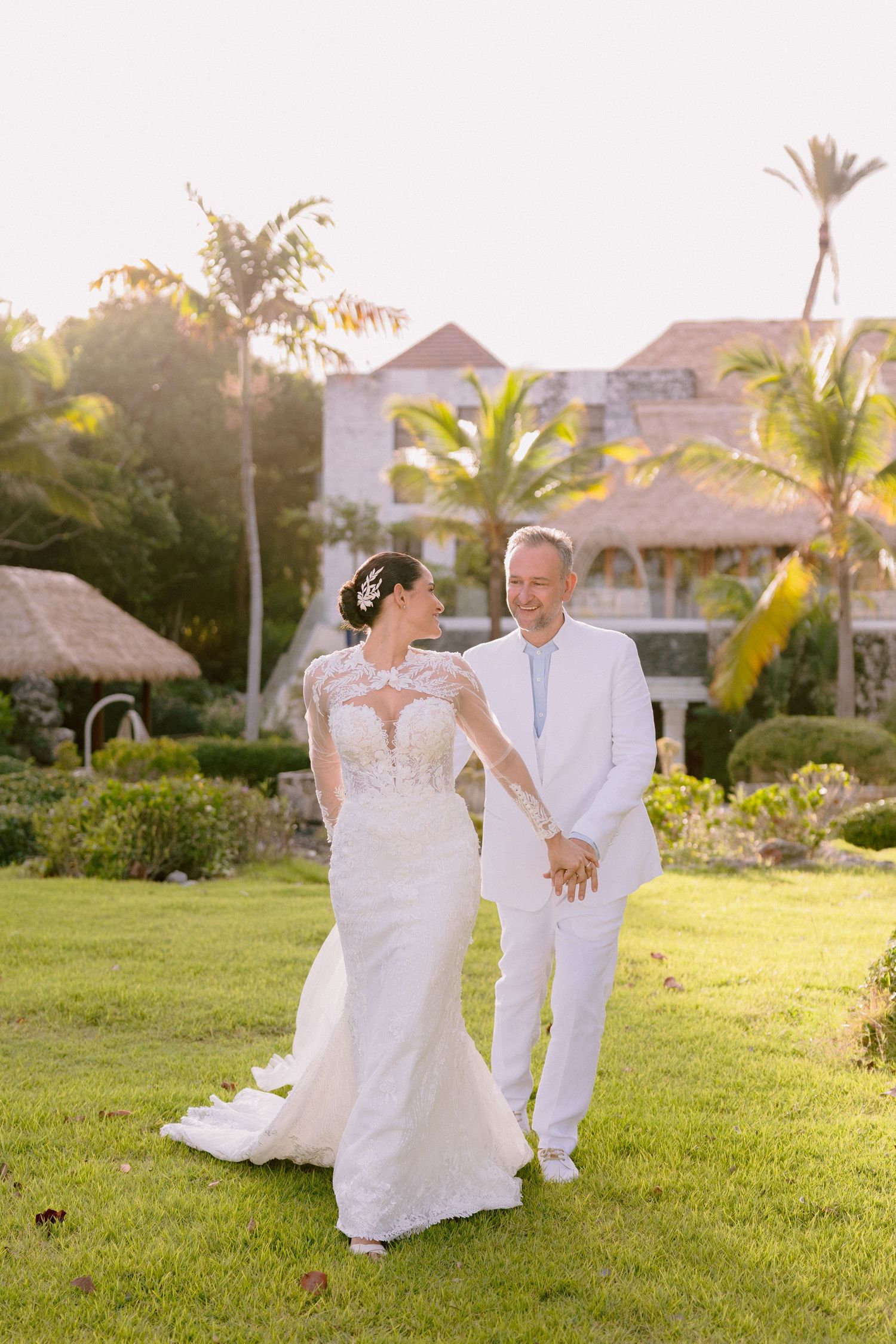 A couple in white attire sits together on lush grass during golden hour at a tropical destination wedding.
