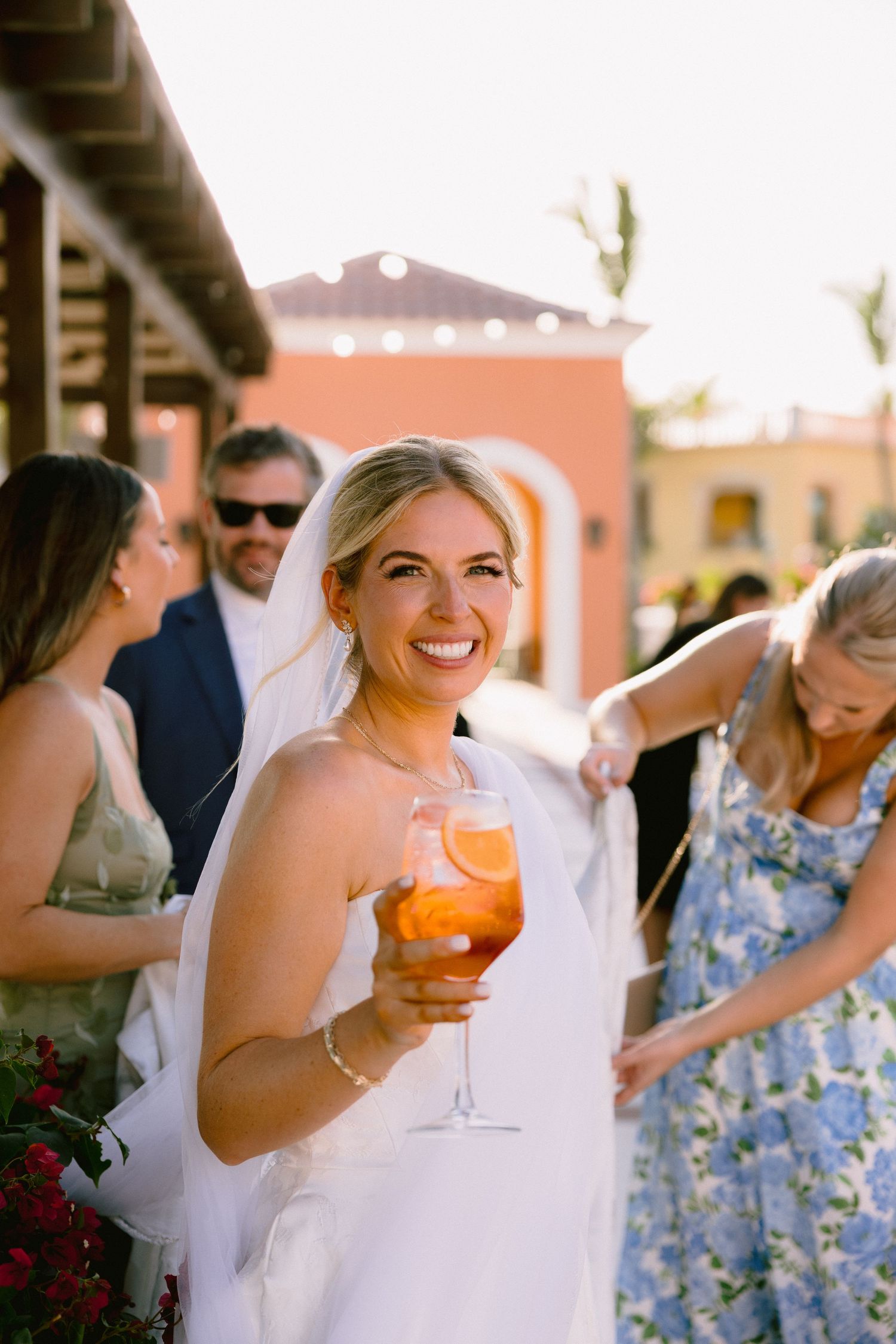 Smiling bride in white dress and veil holds an Aperol Spritz cocktail at an outdoor wedding reception.