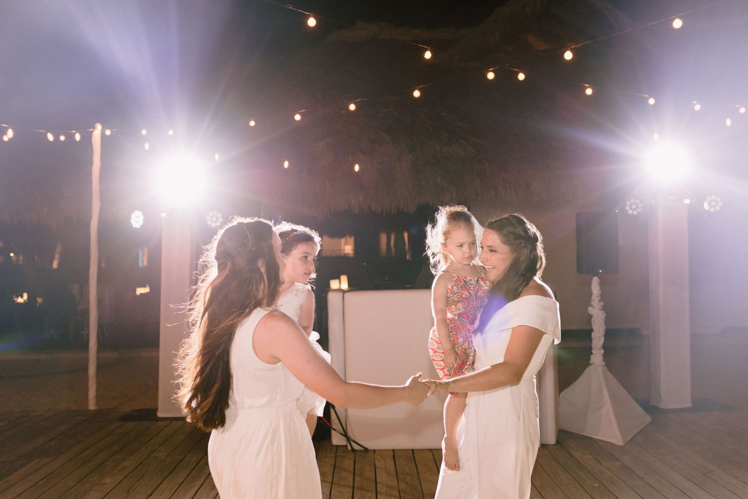 Bridesmaids in white dresses hold hands on a decorated outdoor dance floor with string lights at a wedding reception.