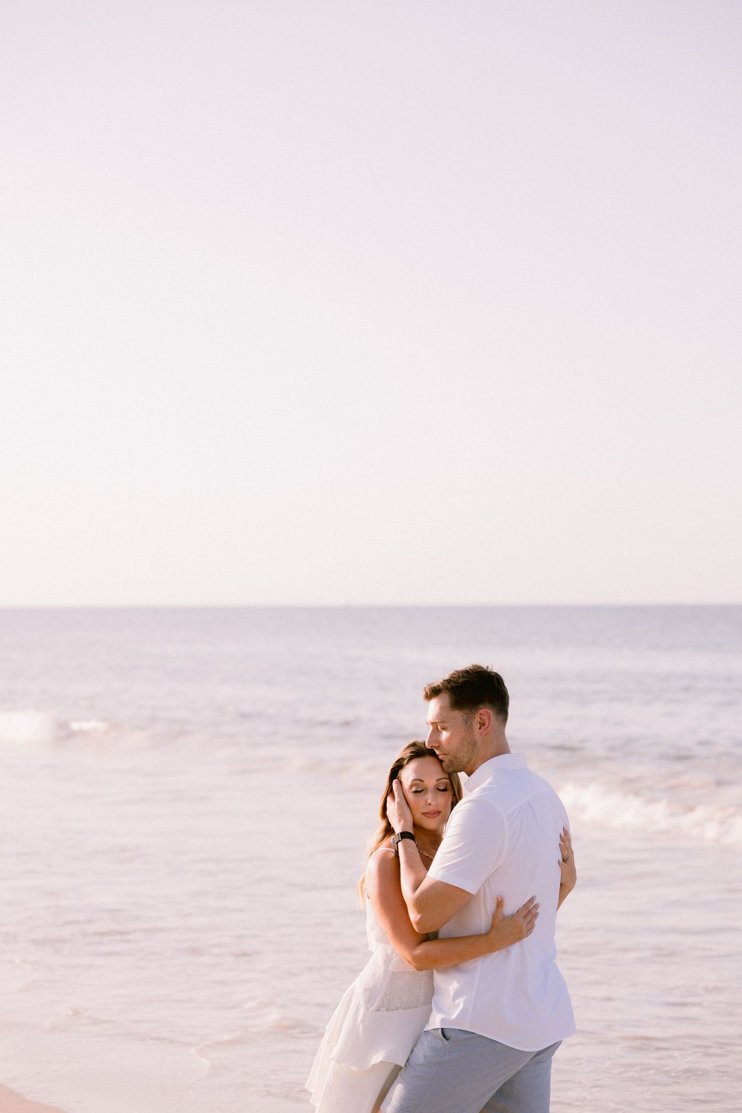 A romantic couple embraces on a serene beach, with gentle ocean waves and a soft pastel sky in the background.