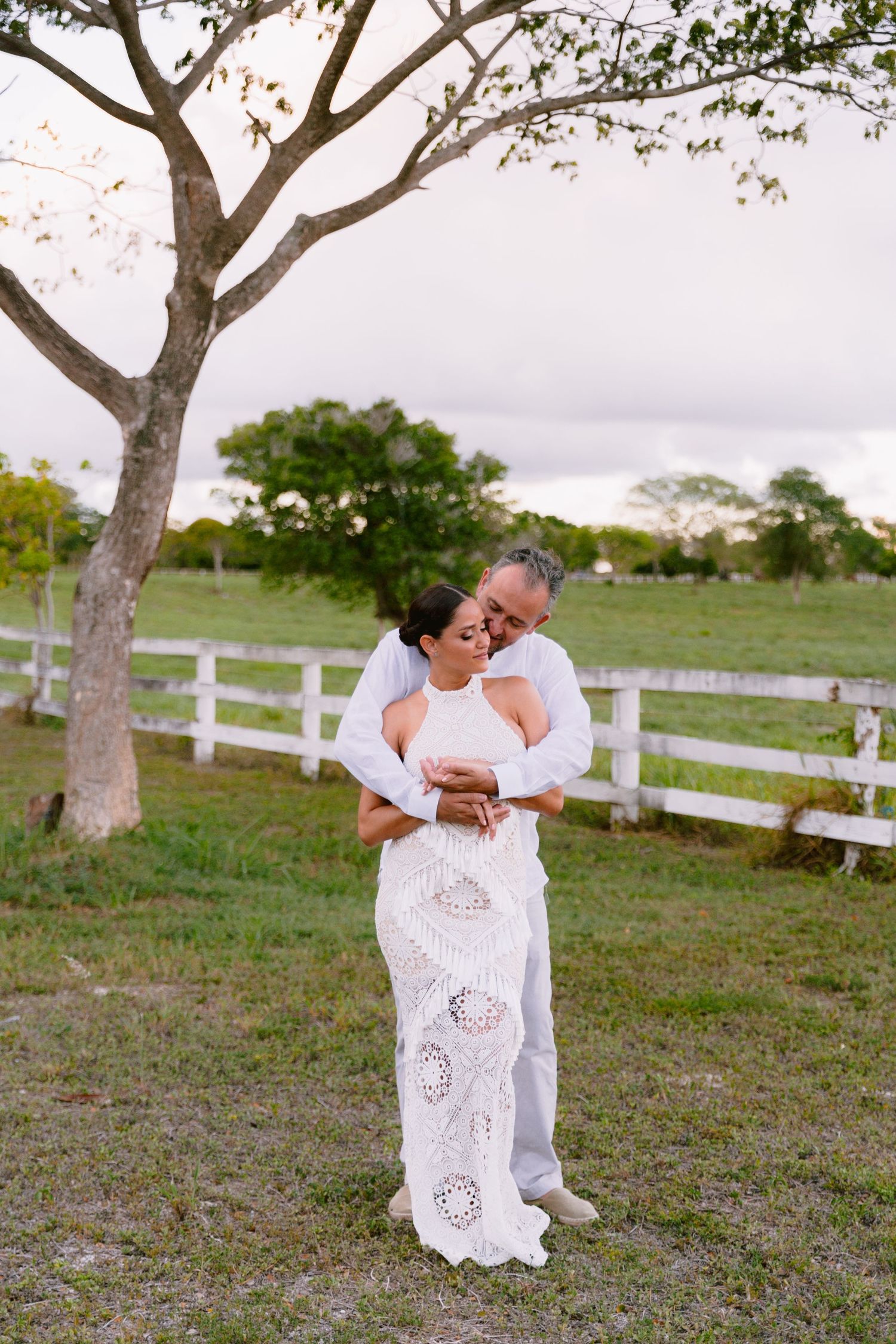 A couple in white attire embraces under a large tree with white fence posts in the background.
