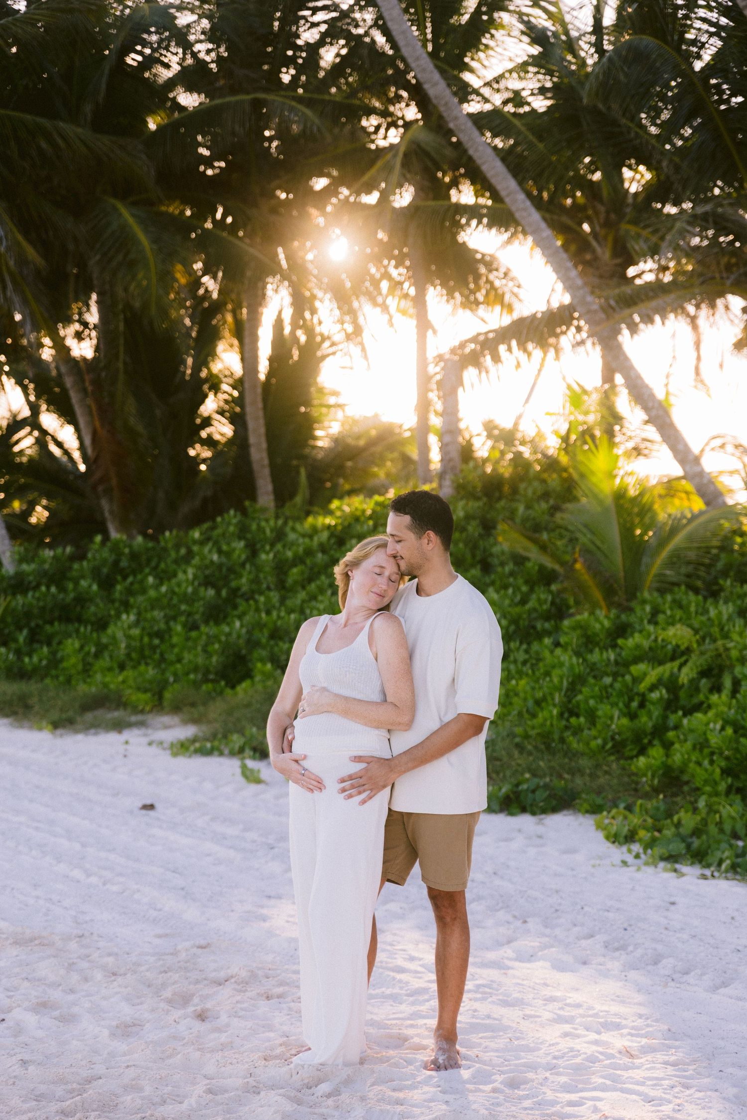 Pregnant couple embracing on a white sand beach at sunset, surrounded by tropical palm trees.