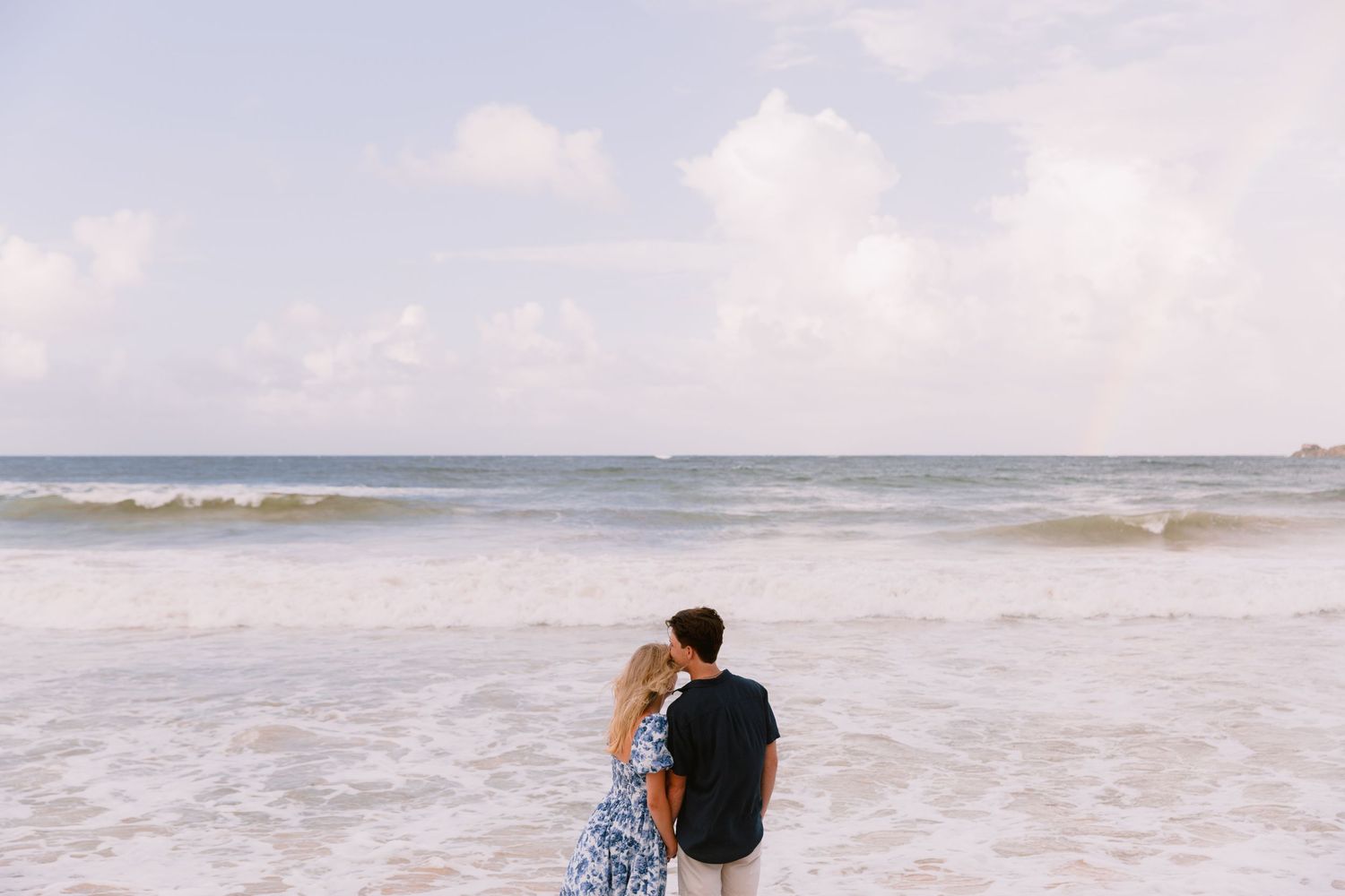 A couple walks along a misty beach shoreline with waves crashing beside them on an overcast day.