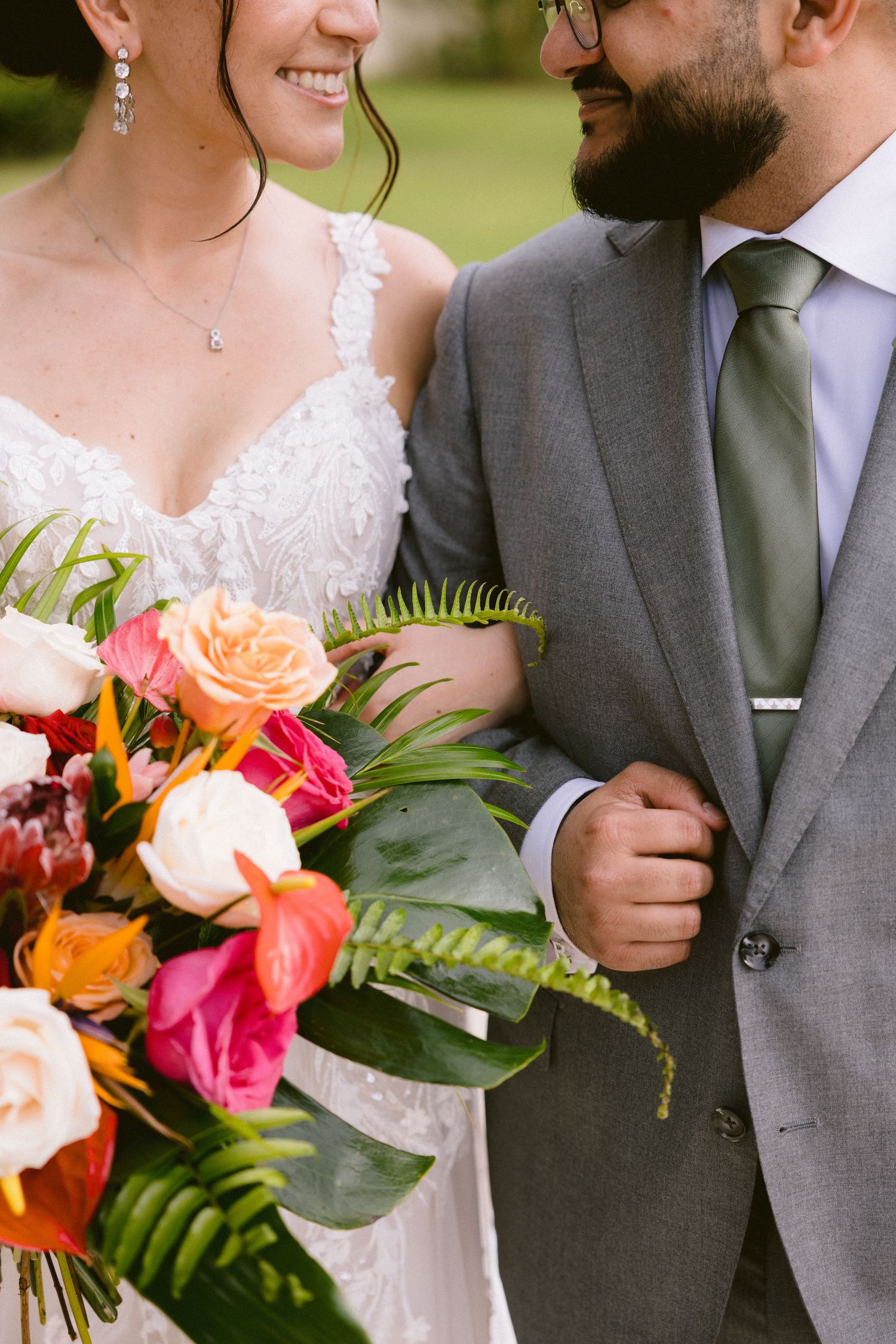 A vibrant tropical wedding bouquet with coral roses and ferns complements a lace wedding dress and gray suit.