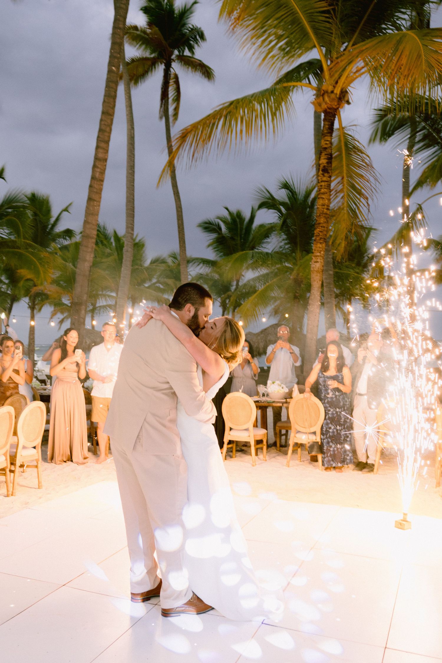 Couple sharing first dance at tropical beach wedding with sparklers and palm trees under evening sky.