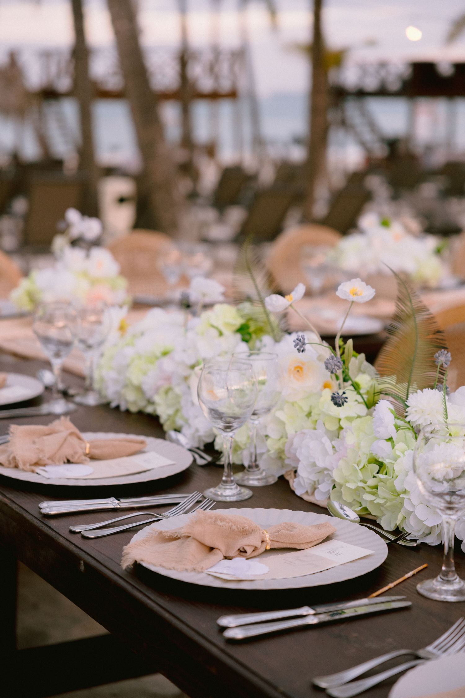 Elegant outdoor wedding reception table with white floral centerpieces, champagne napkins, and crystal glassware by the sea.