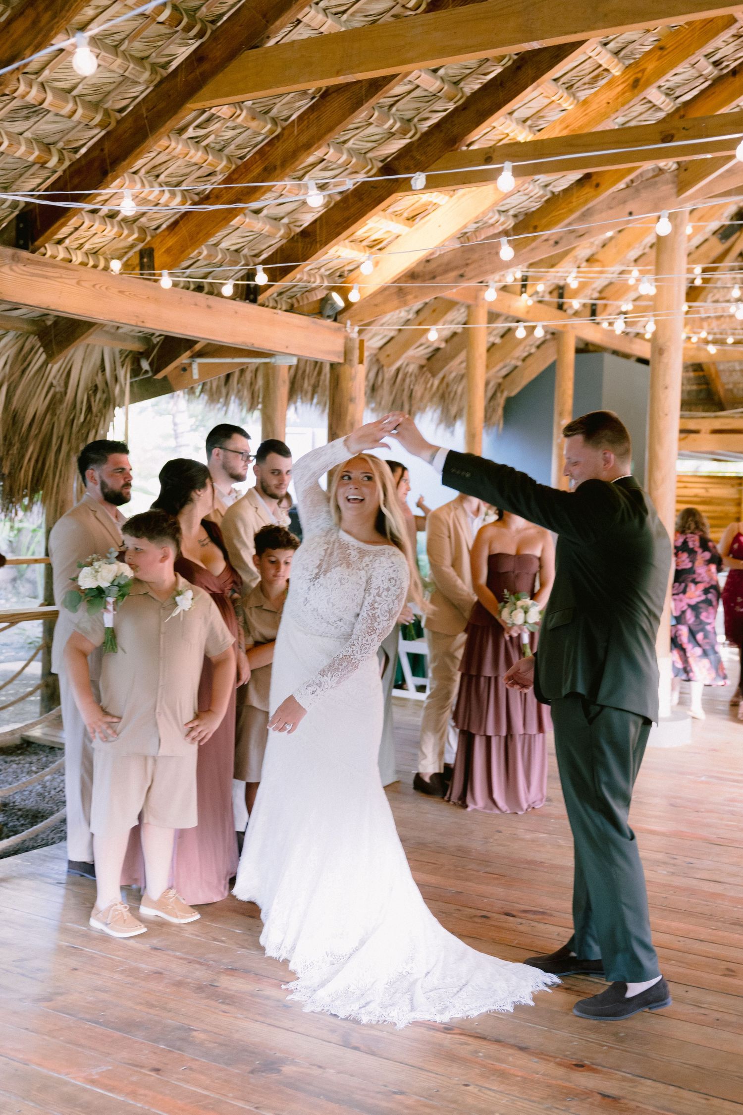 Groom twirls bride in white lace gown during wedding ceremony in rustic barn venue with string lights.