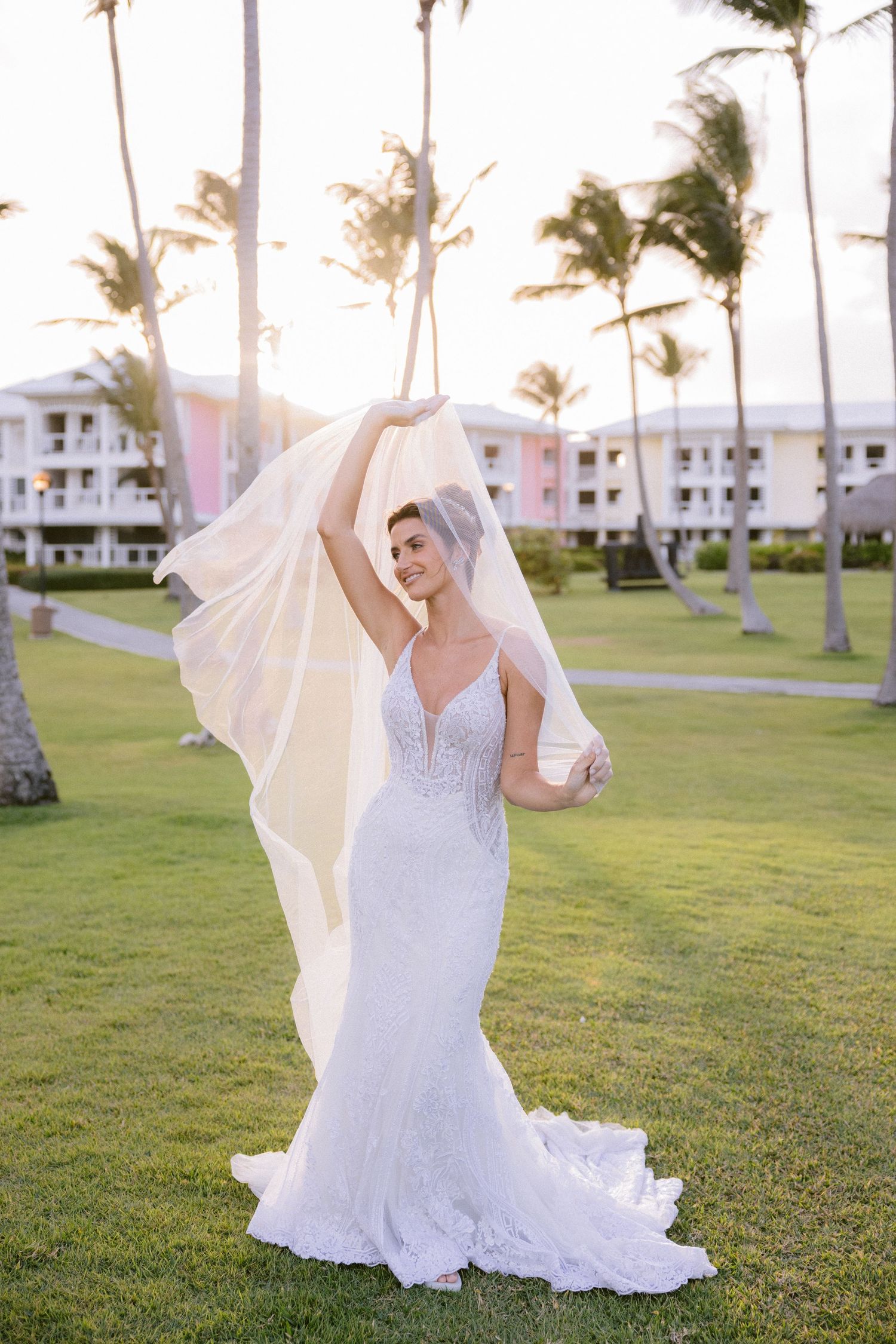 Bride in lace mermaid wedding gown lifting veil outdoors at tropical resort with palm trees at golden hour.
