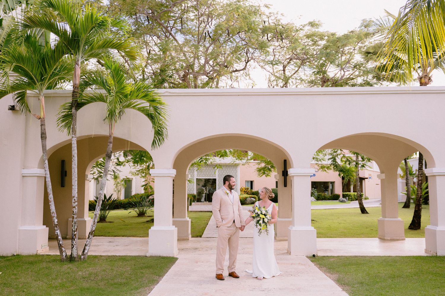 Bride and groom stand under elegant arched colonnade surrounded by palm trees at a tropical wedding venue.