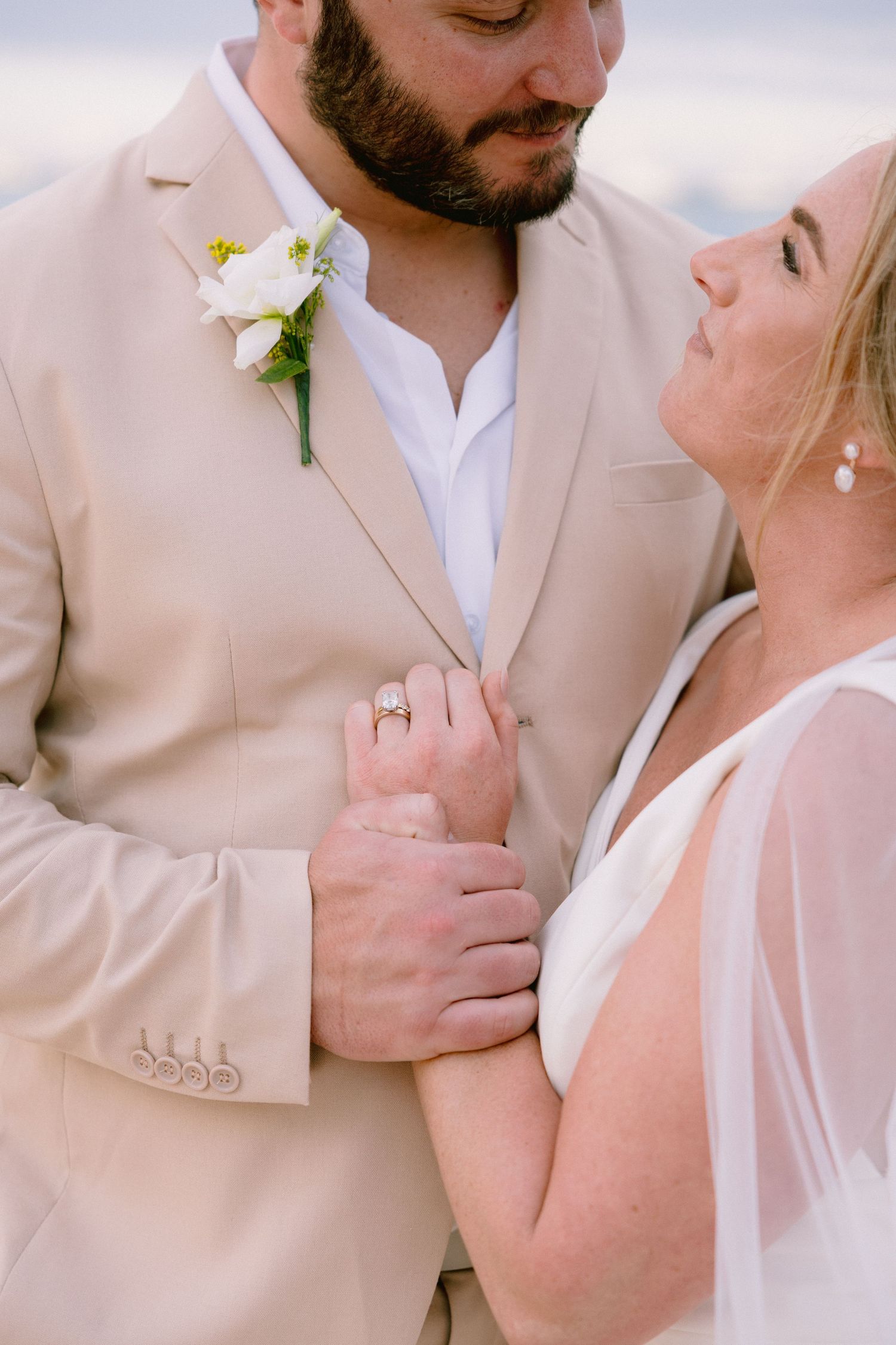 Bride and groom in beige suit with white boutonniere share a tender moment, bride wearing diamond ring and white gown.
