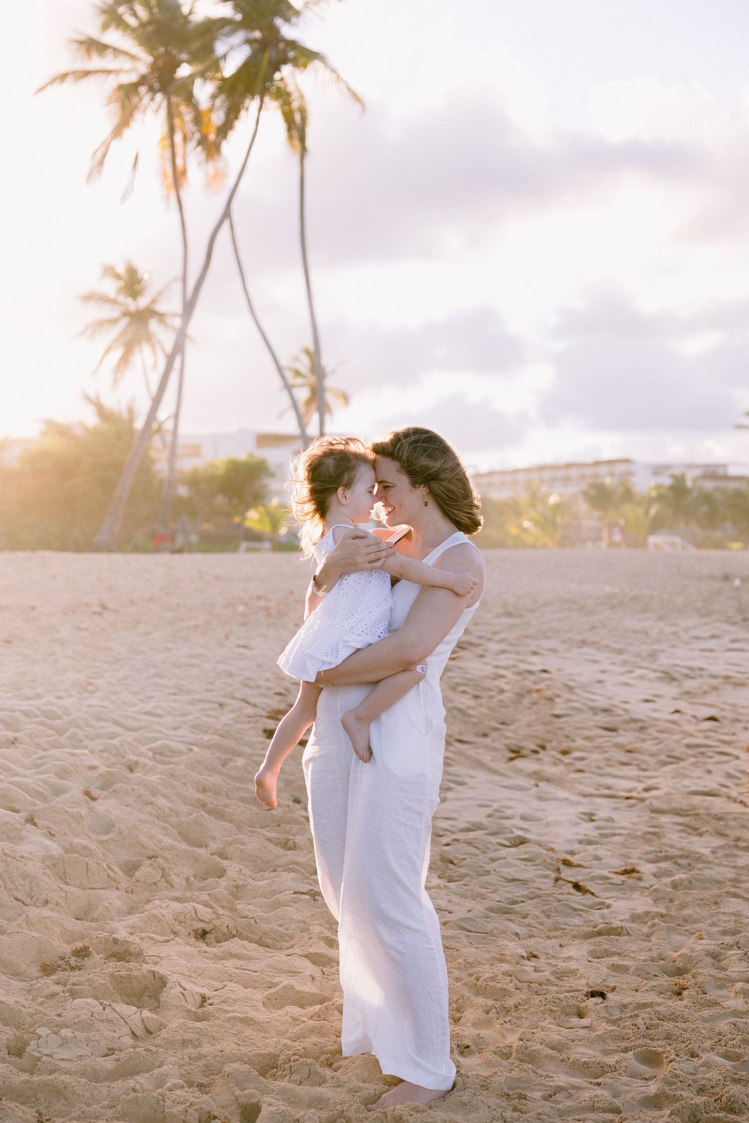 A mother holds her child on a sandy beach at golden sunset, with palm trees and soft pastel sky behind them.