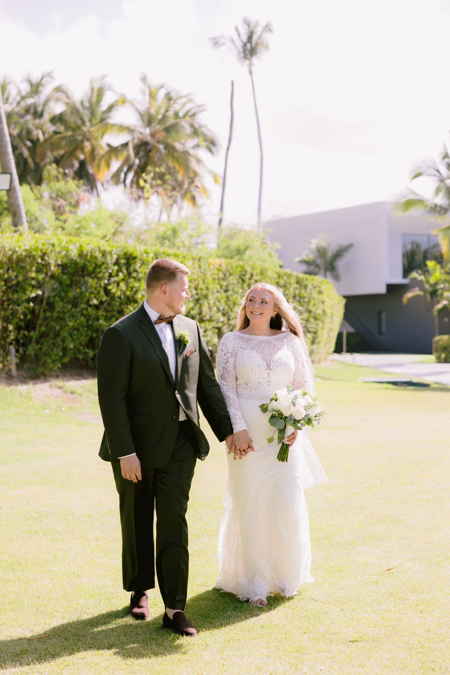 Bride and groom walk hand in hand on green lawn with palm trees, she holds white bouquet, he wears black tuxedo.