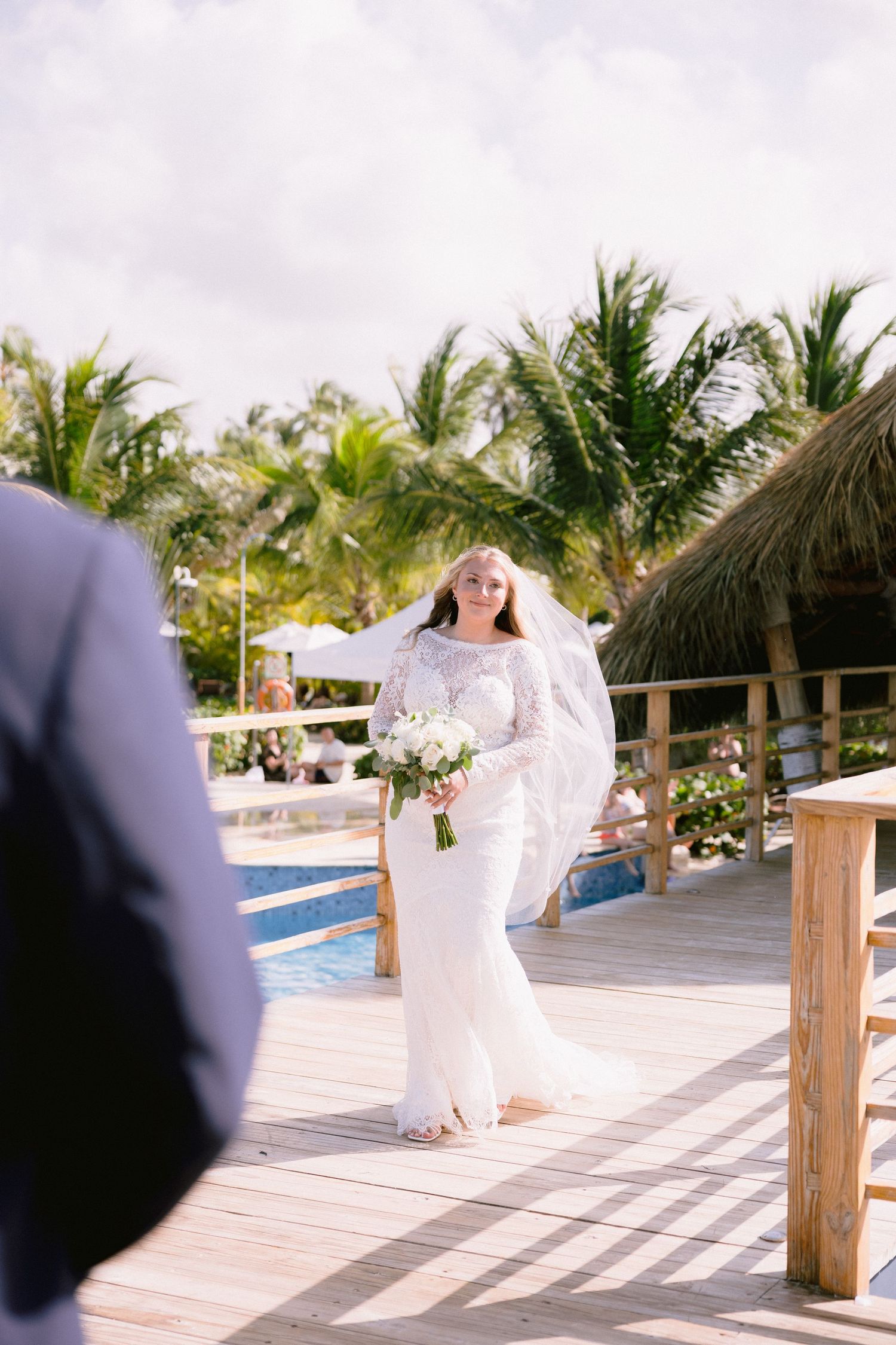 Bride in white gown and veil walks along tropical resort deck holding bouquet, with palm trees and pool backdrop.