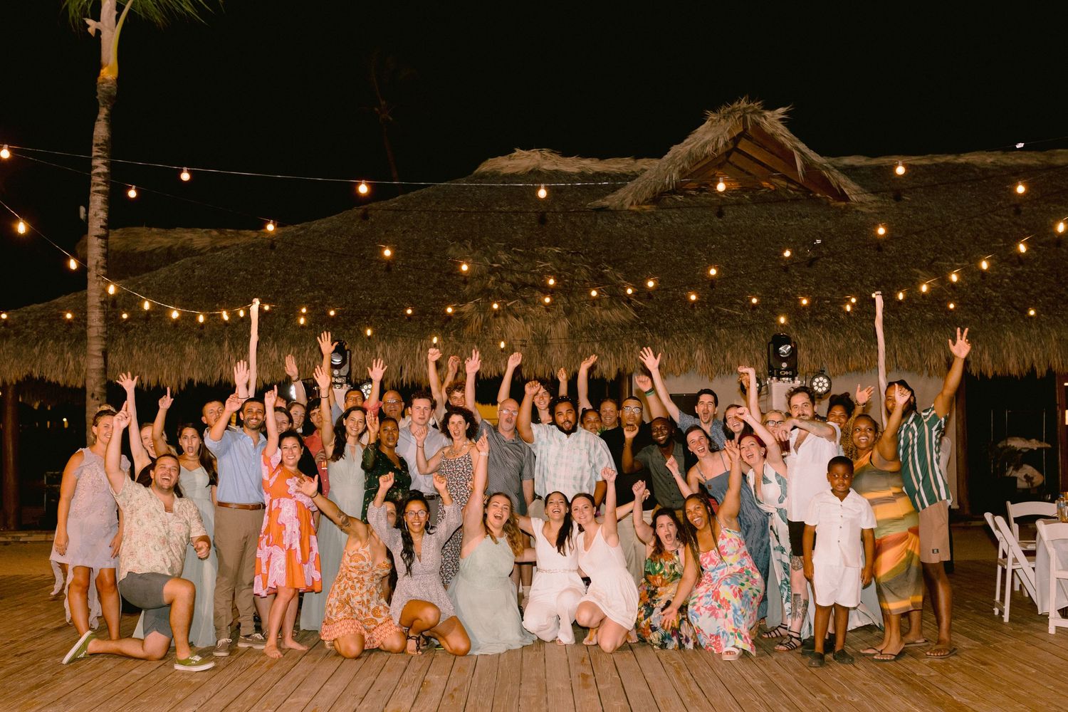 Large group celebrating at an outdoor tropical wedding reception with string lights and thatched roof at night.