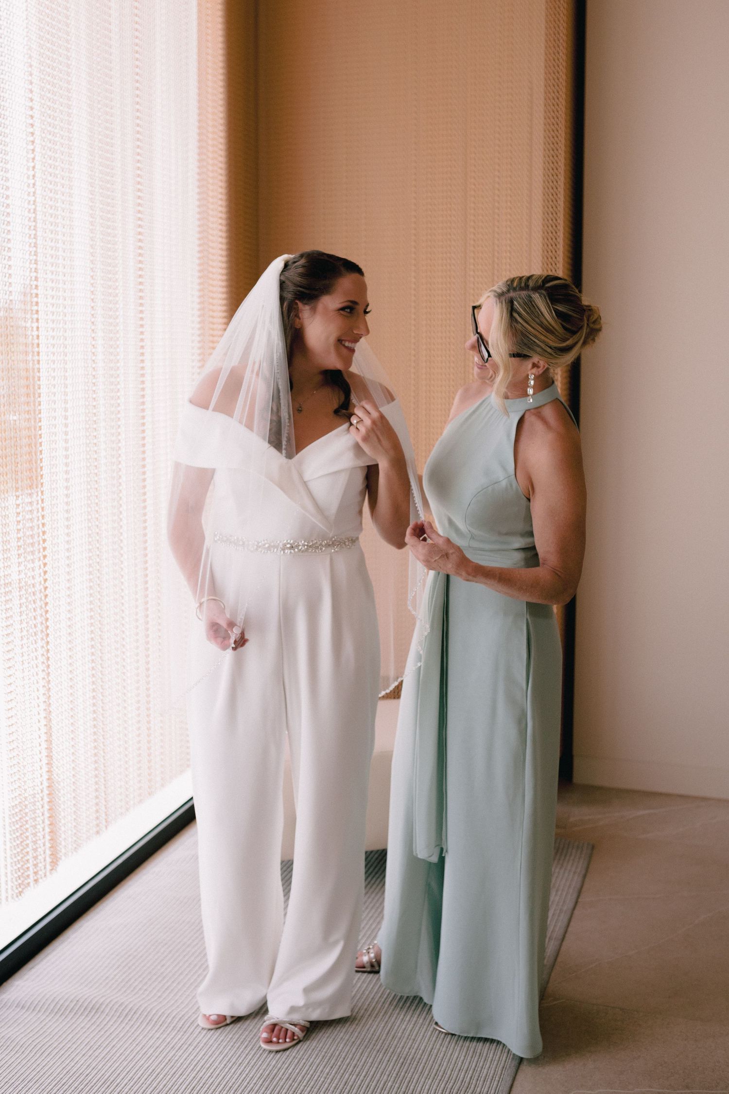 Bride in white jumpsuit and veil smiling with bridesmaid in mint green gown near window.