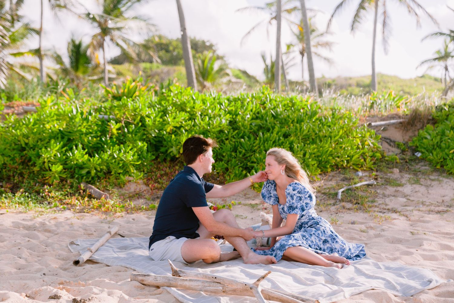 A couple shares a romantic moment on a sandy beach with palm trees in the background during sunset.