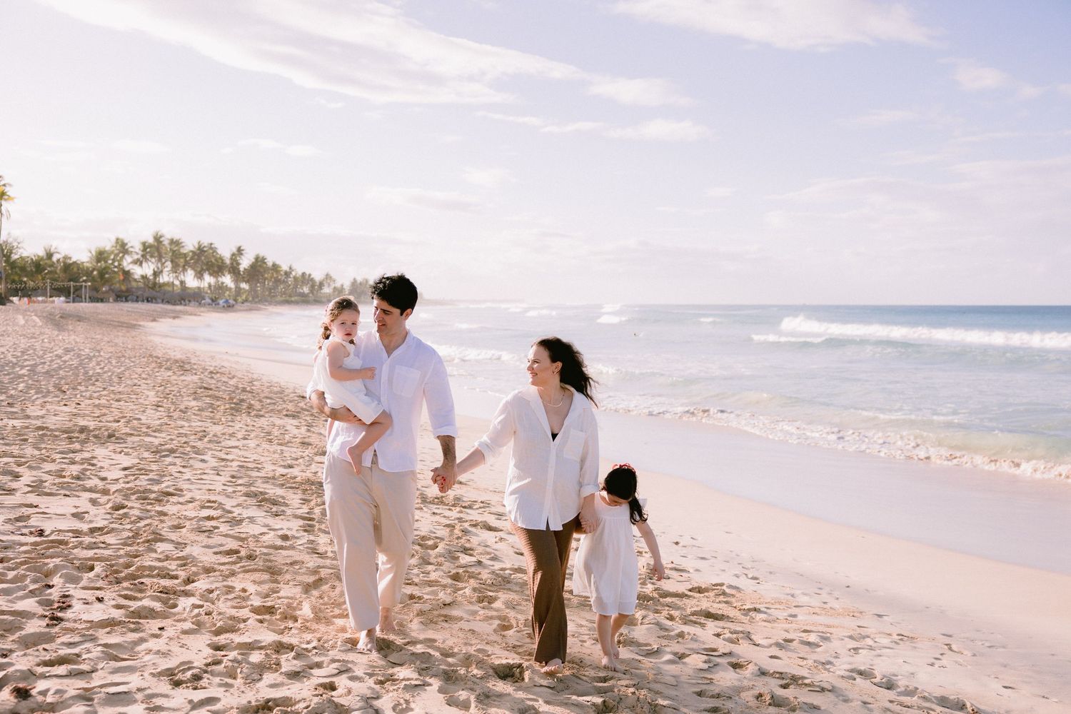 A family walks together along a scenic tropical beach with palm trees and ocean waves in the background.