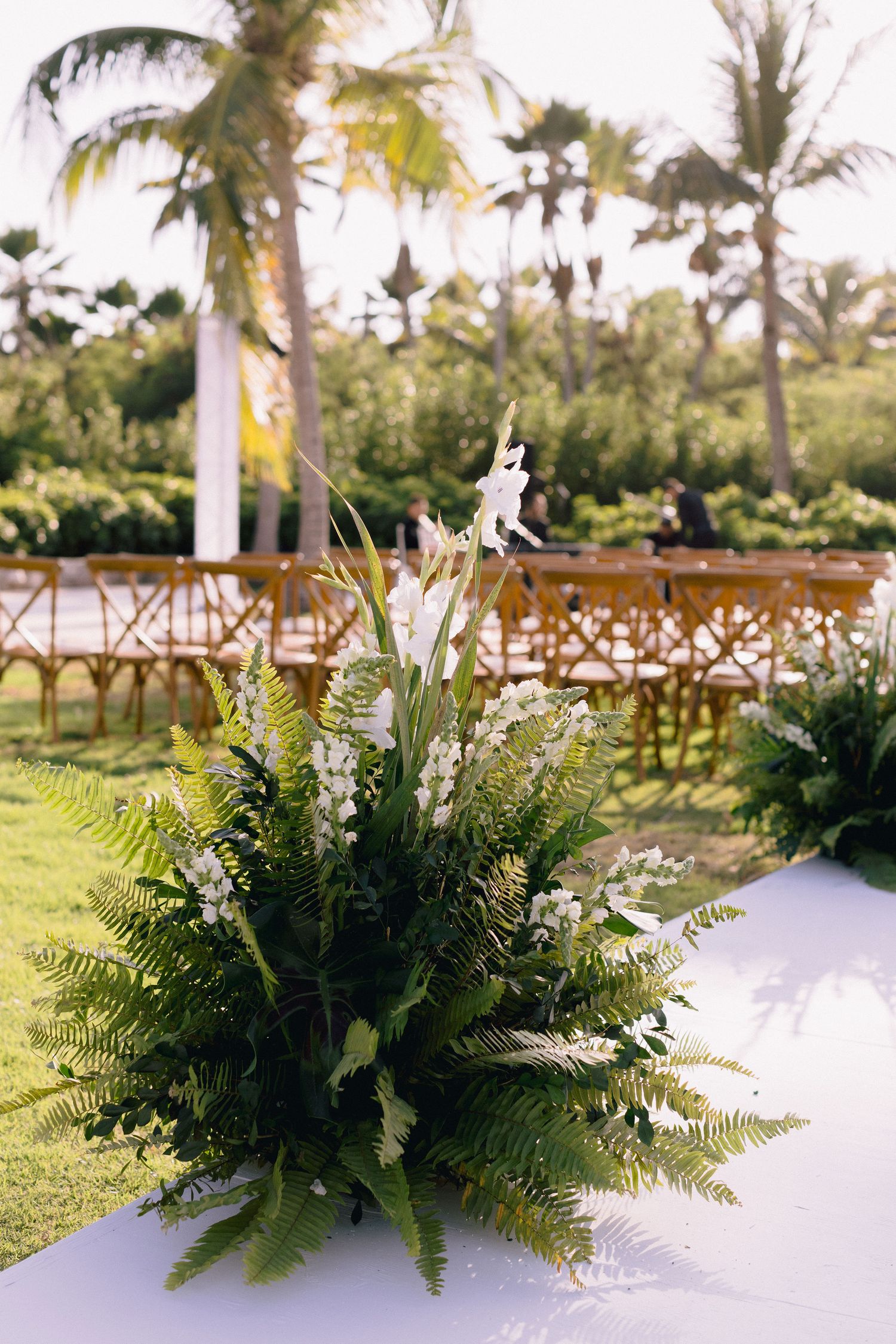 Lush green fern and white floral arrangements decorate outdoor tables at a tropical wedding venue with palm trees.