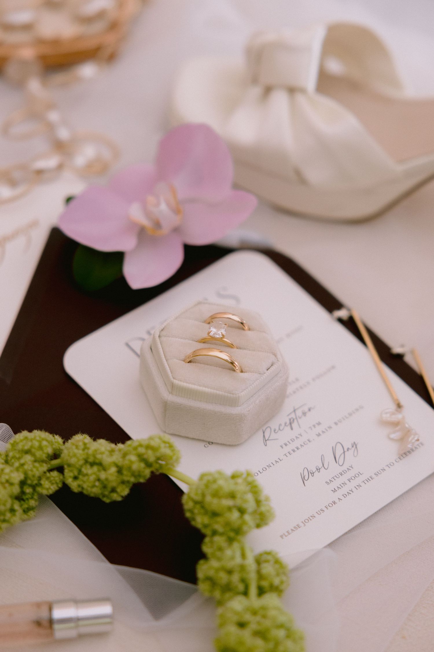 Gold wedding rings in a velvet box on wedding invitation, with orchid, green florals, and bridal heel in background.