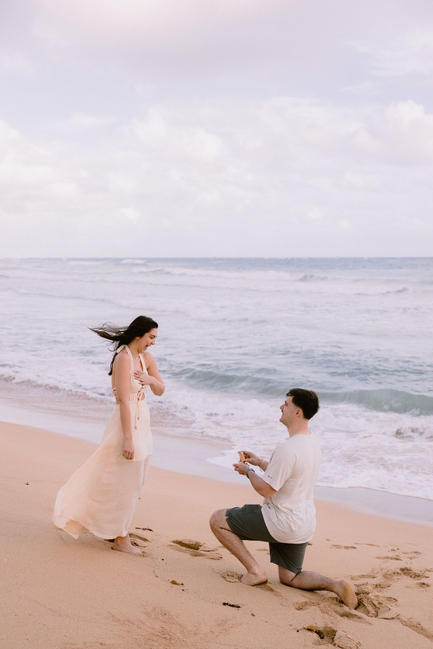Man proposes to woman on sandy beach with ocean waves, she covers her mouth in surprise wearing a pink dress.