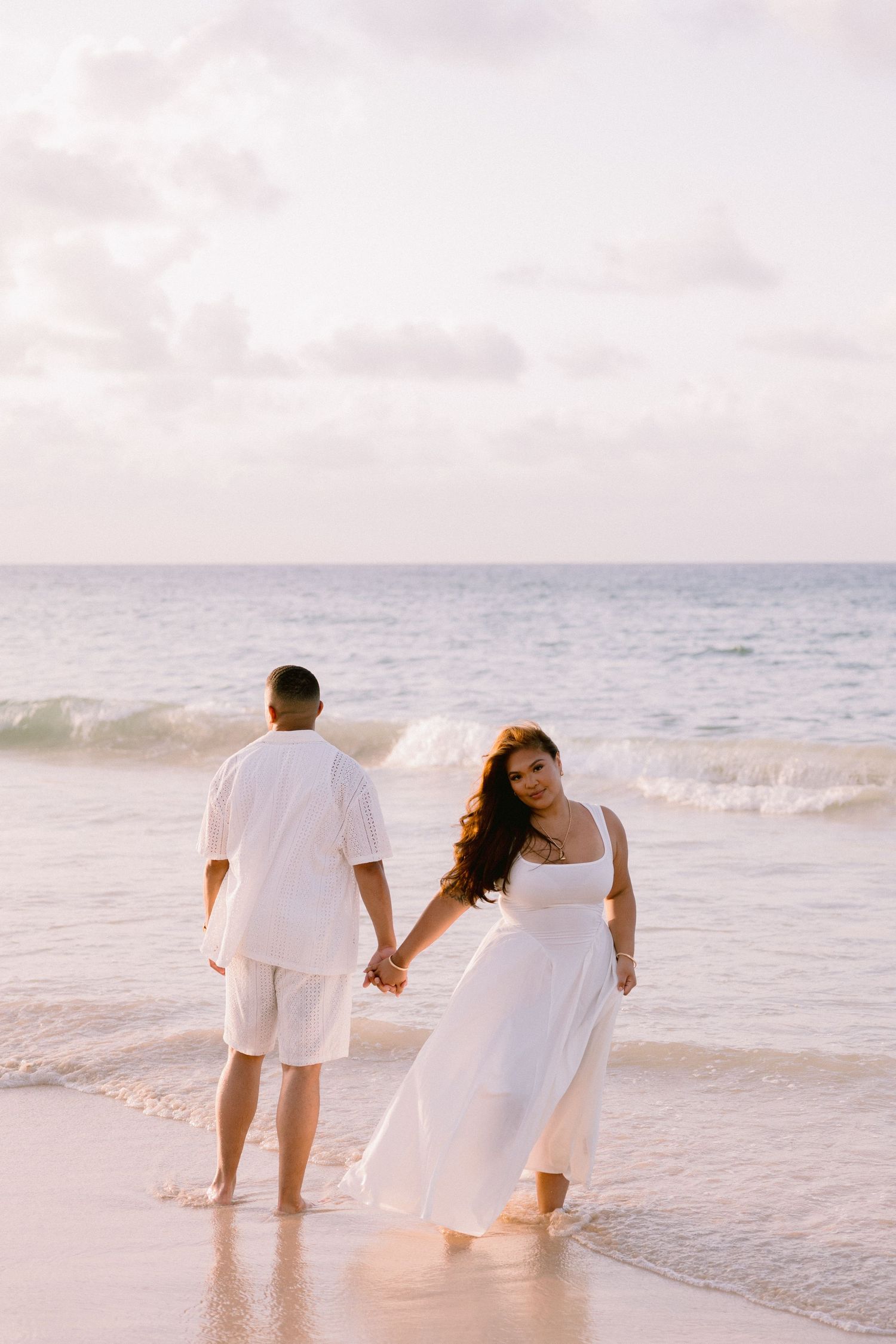 Couple in white outfits holding hands on a sandy beach at sunset, woman in flowing dress looking back at camera.