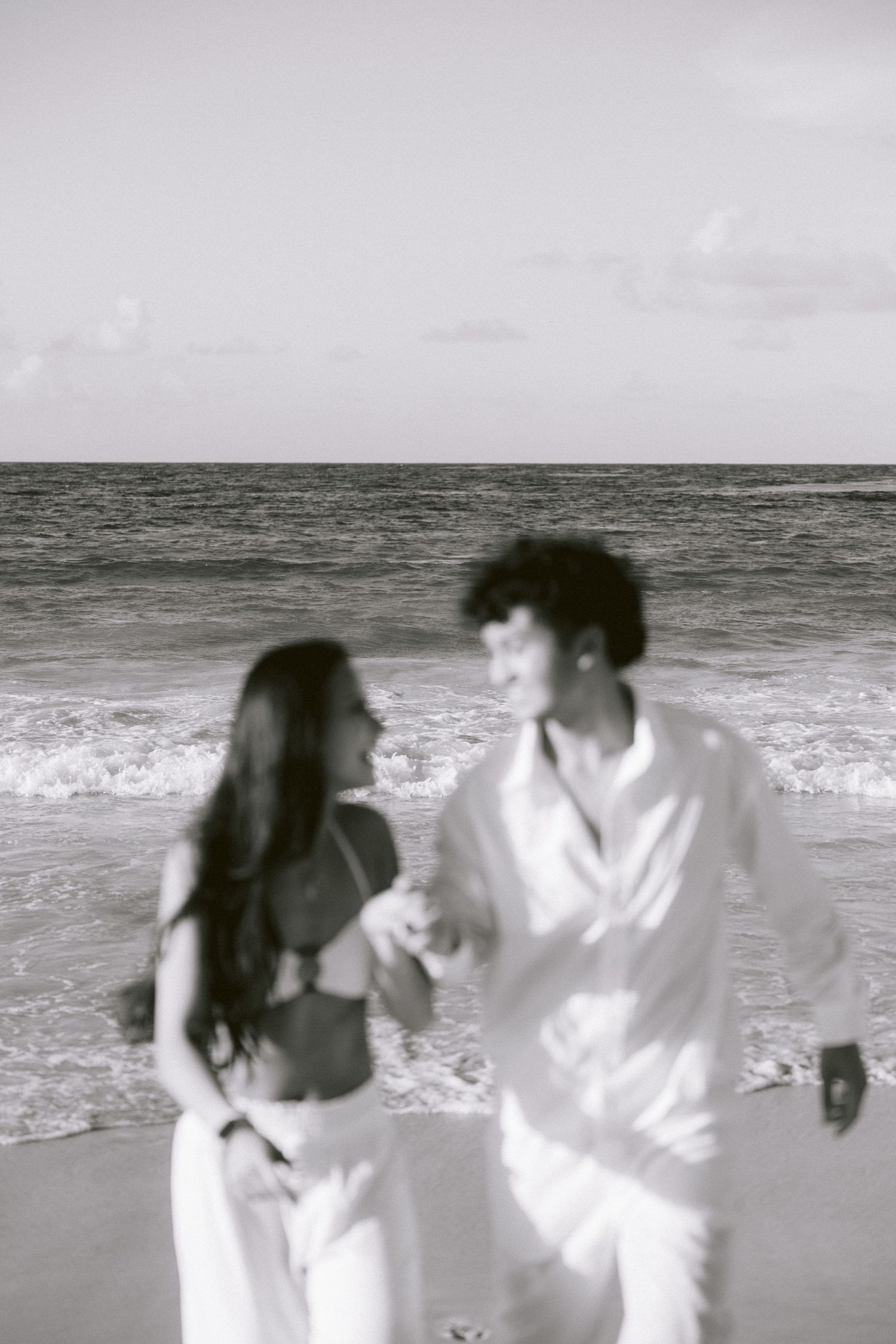 A couple in white attire running playfully along the beach shore.