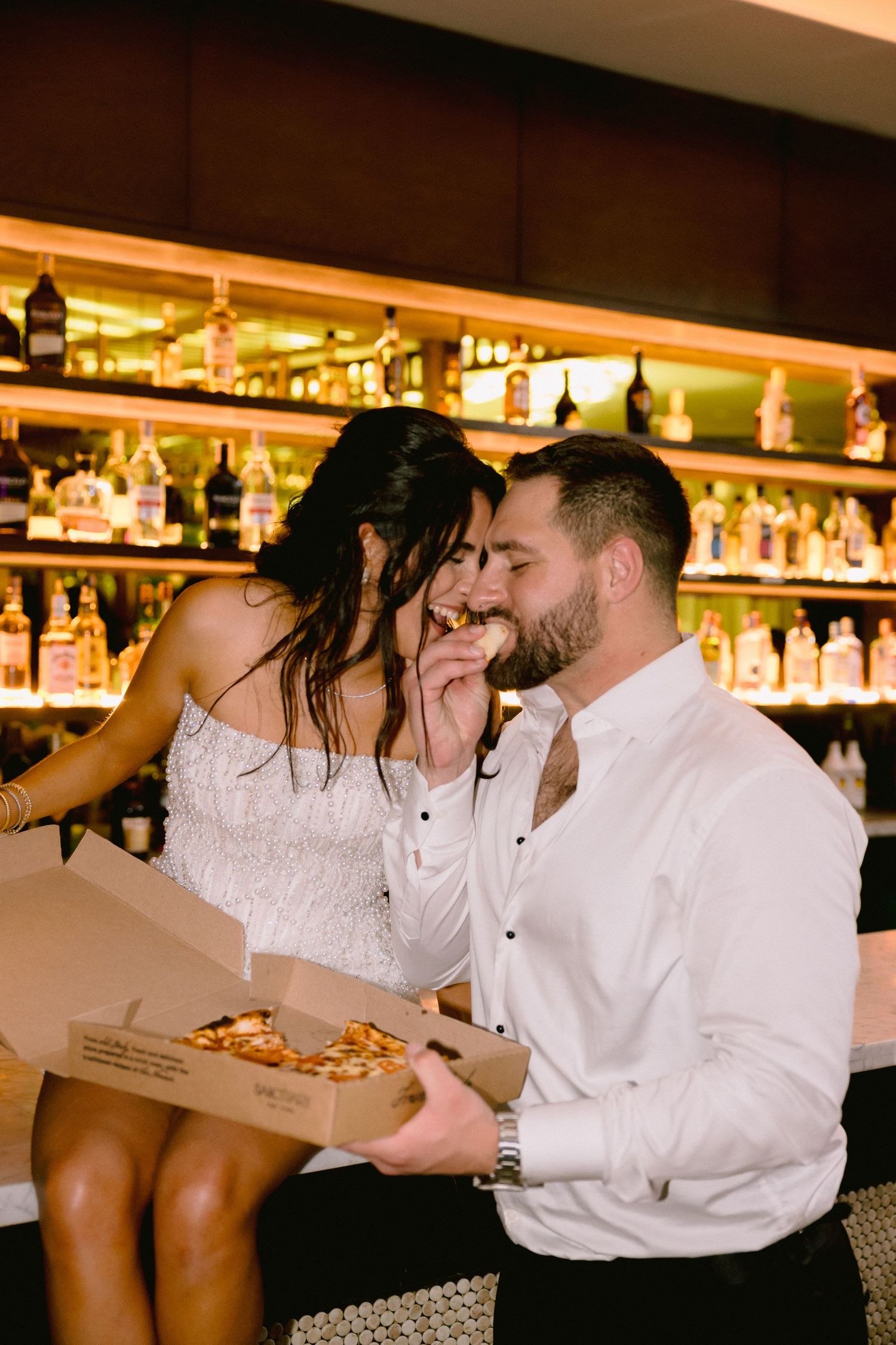 Bride and groom sharing pizza at a bar with backlit liquor bottles, celebrating their wedding reception.