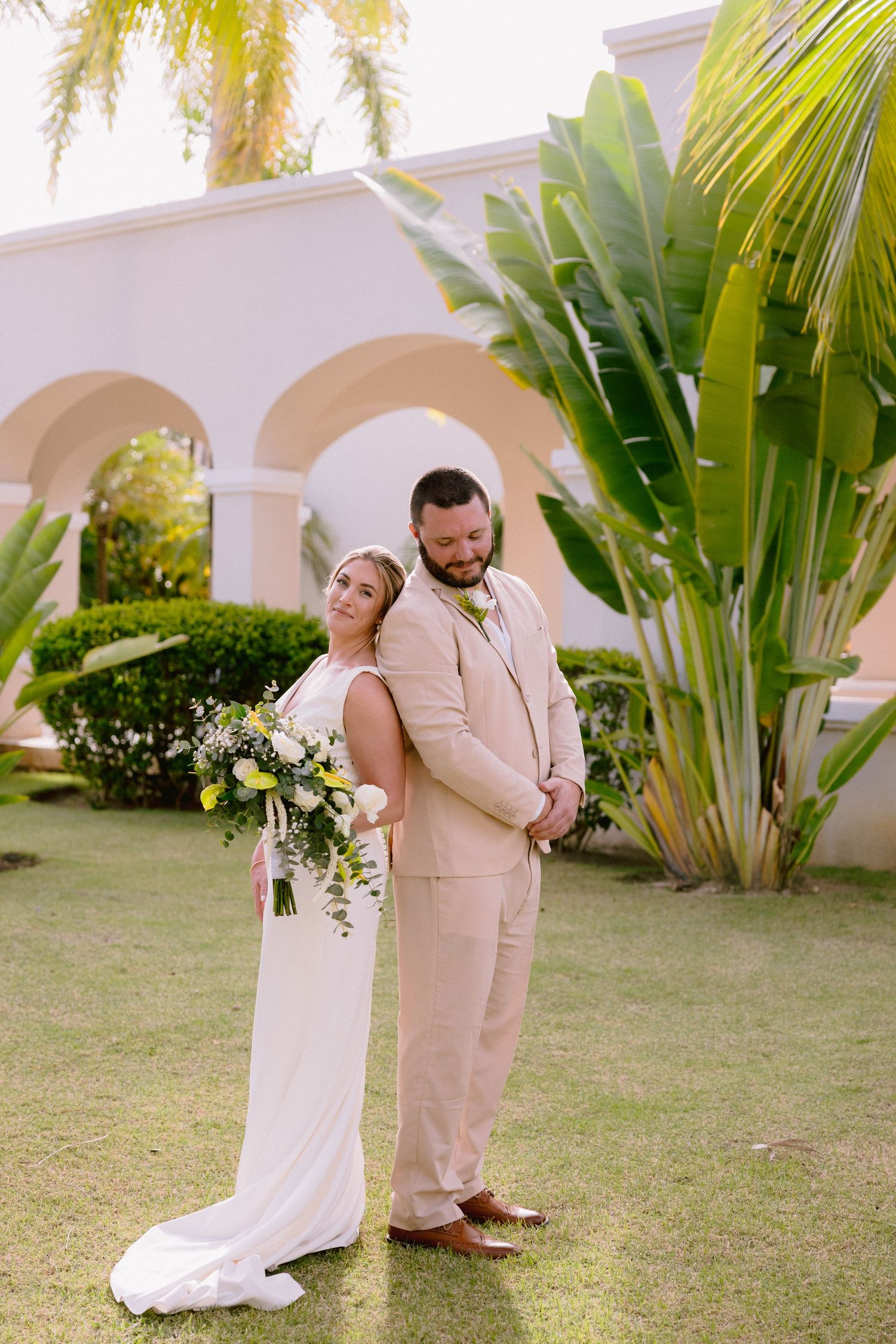 Bride and groom pose back-to-back in tropical garden, she holds floral bouquet, he wears tan suit, white building behind them.