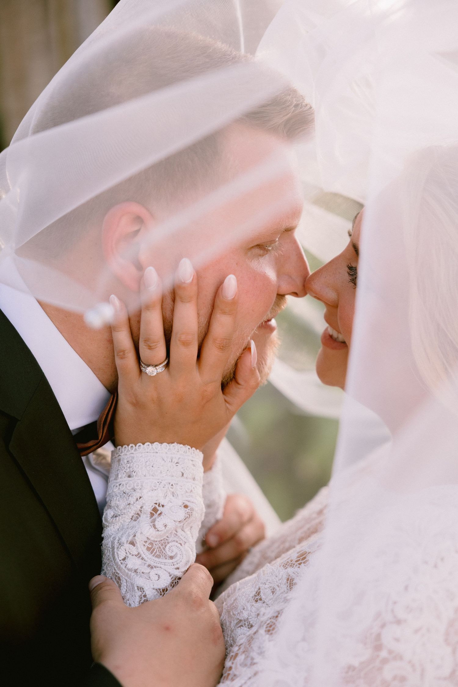 Bride and groom share an intimate moment under a flowing veil, showcasing a diamond engagement ring on her lace sleeve.