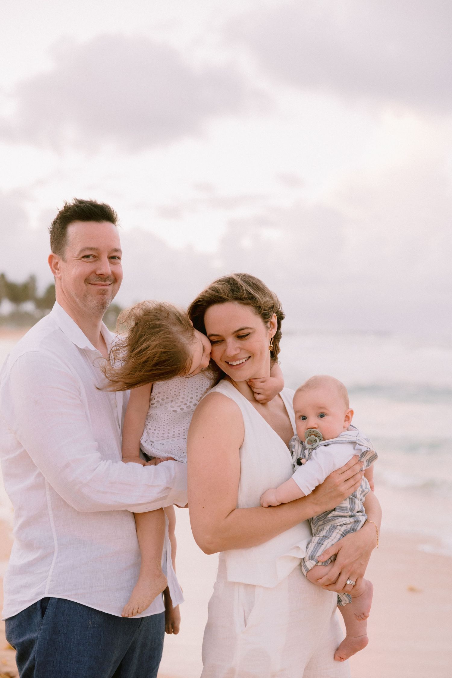 Family of four poses on a beach at sunset, parents holding two young children, dressed in white and casual attire.