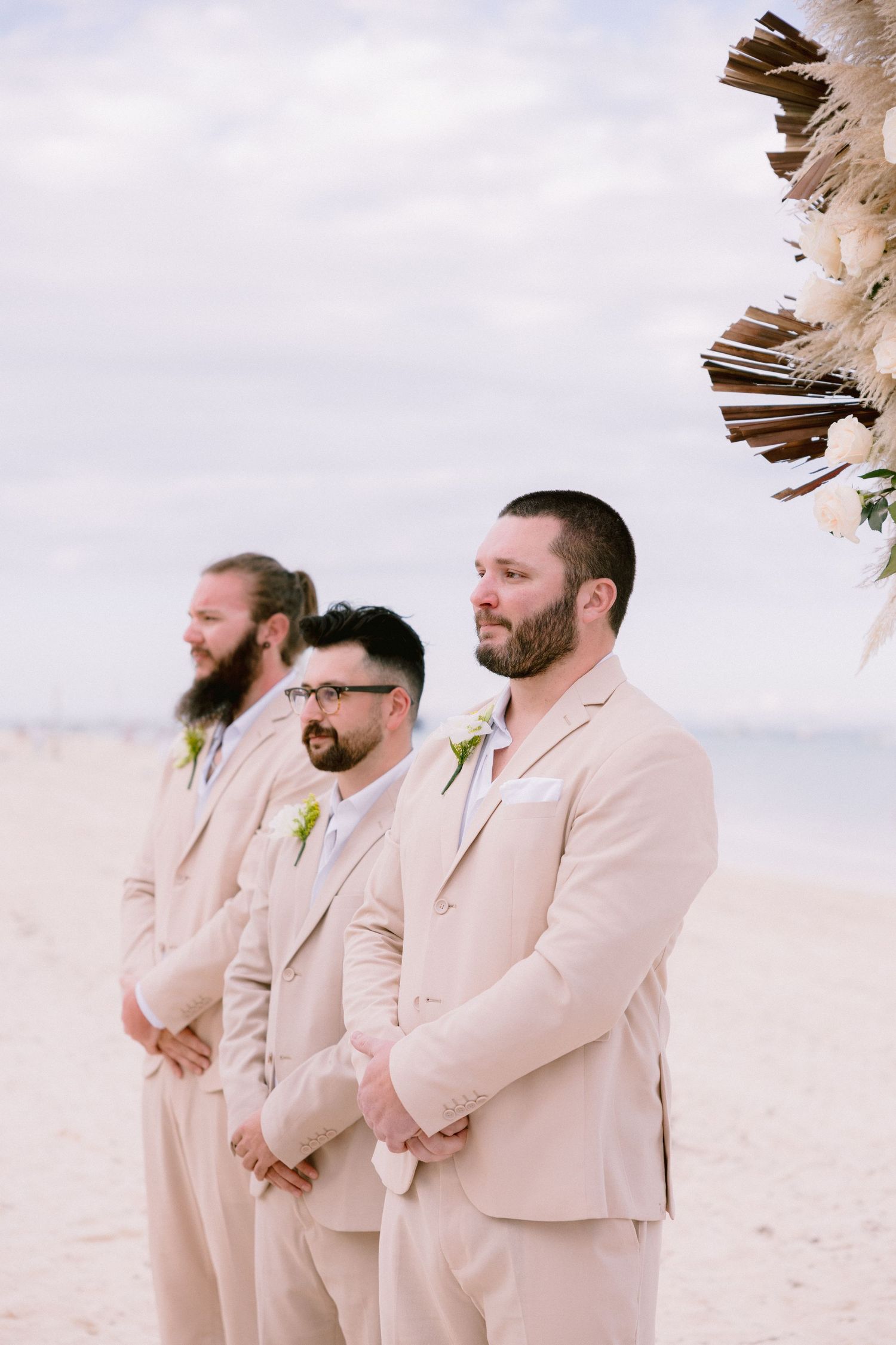 Three groomsmen in matching beige suits with boutonnieres standing on a beach during an outdoor wedding ceremony.