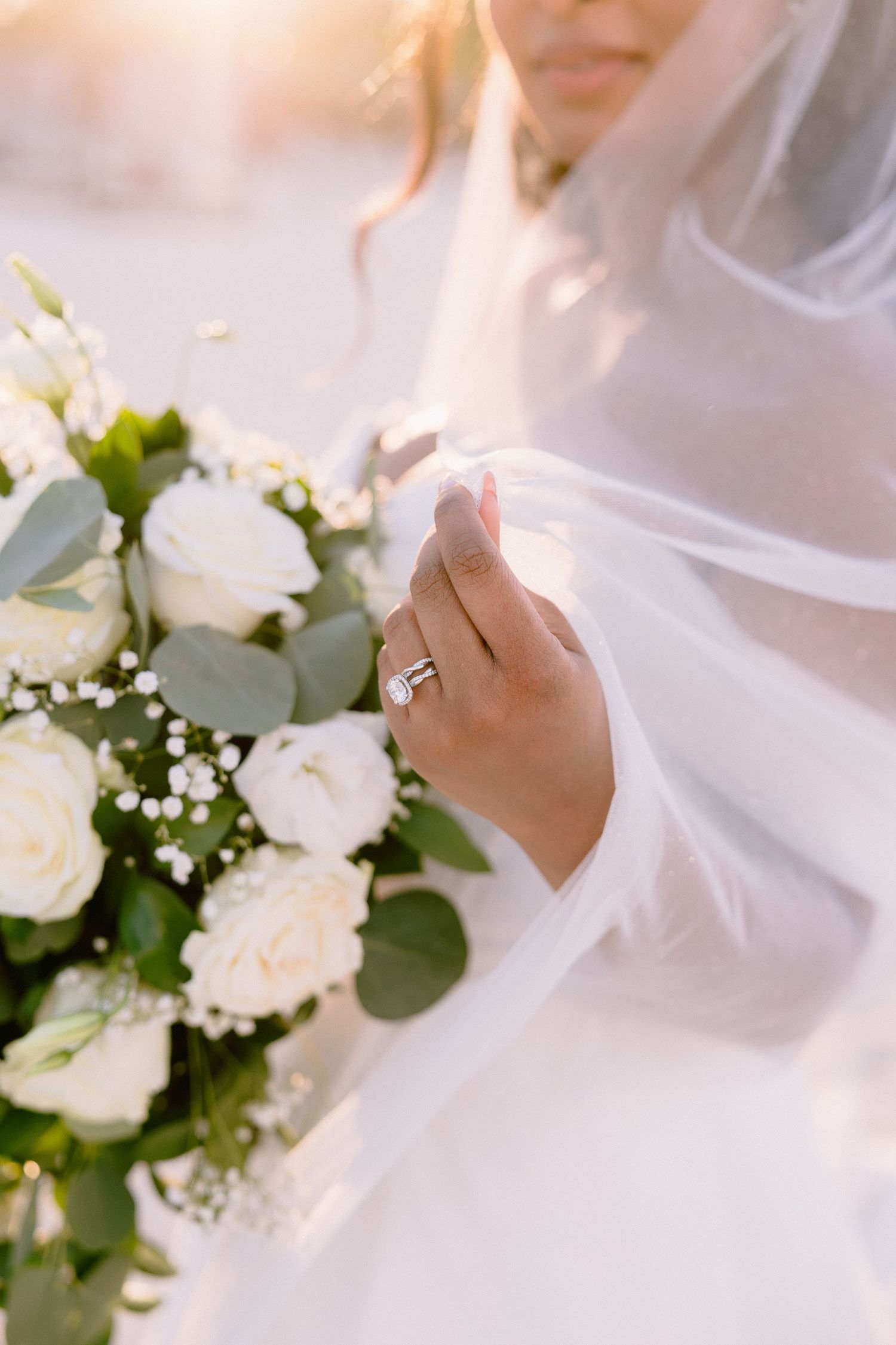 Bride holding white rose bouquet with eucalyptus, wearing diamond engagement ring, soft veil draped over hand in golden light.