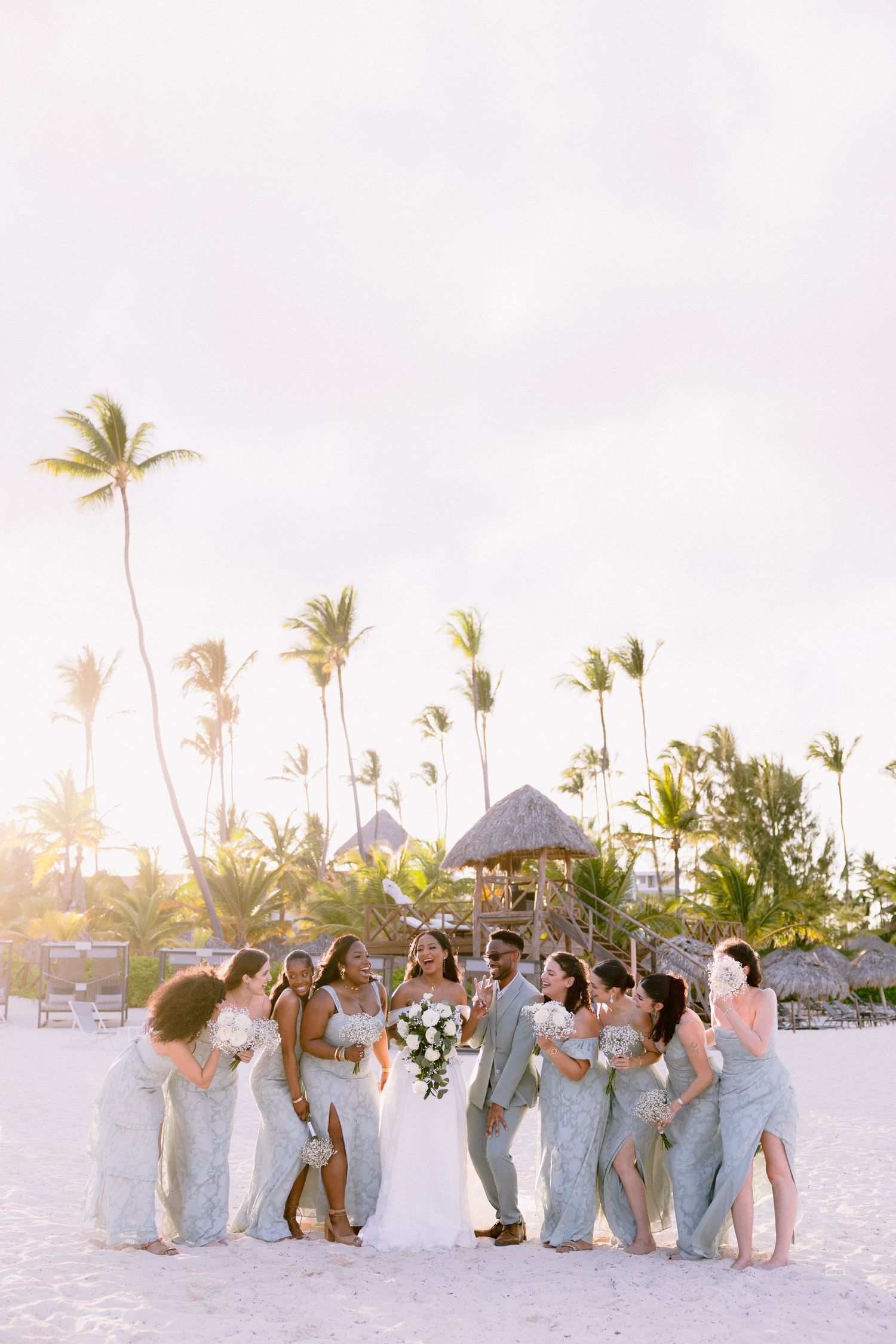 Bride and bridesmaids in blue gowns celebrate on a tropical beach with palm trees at golden hour.