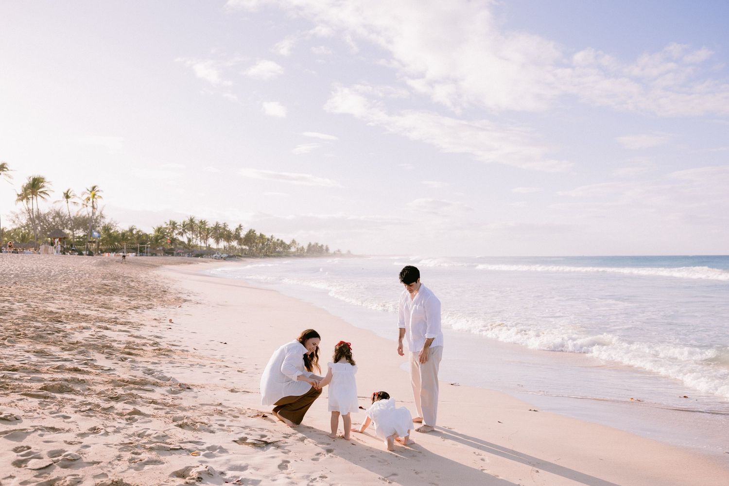 A couple in white attire walks along a tropical beach shoreline with palm trees and ocean waves in the background.