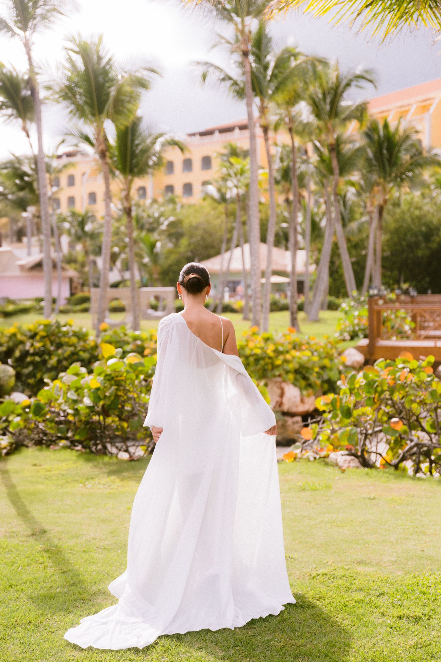 Bride in elegant white one-shoulder gown with flowing train stands on lush green lawn surrounded by tropical palm trees.