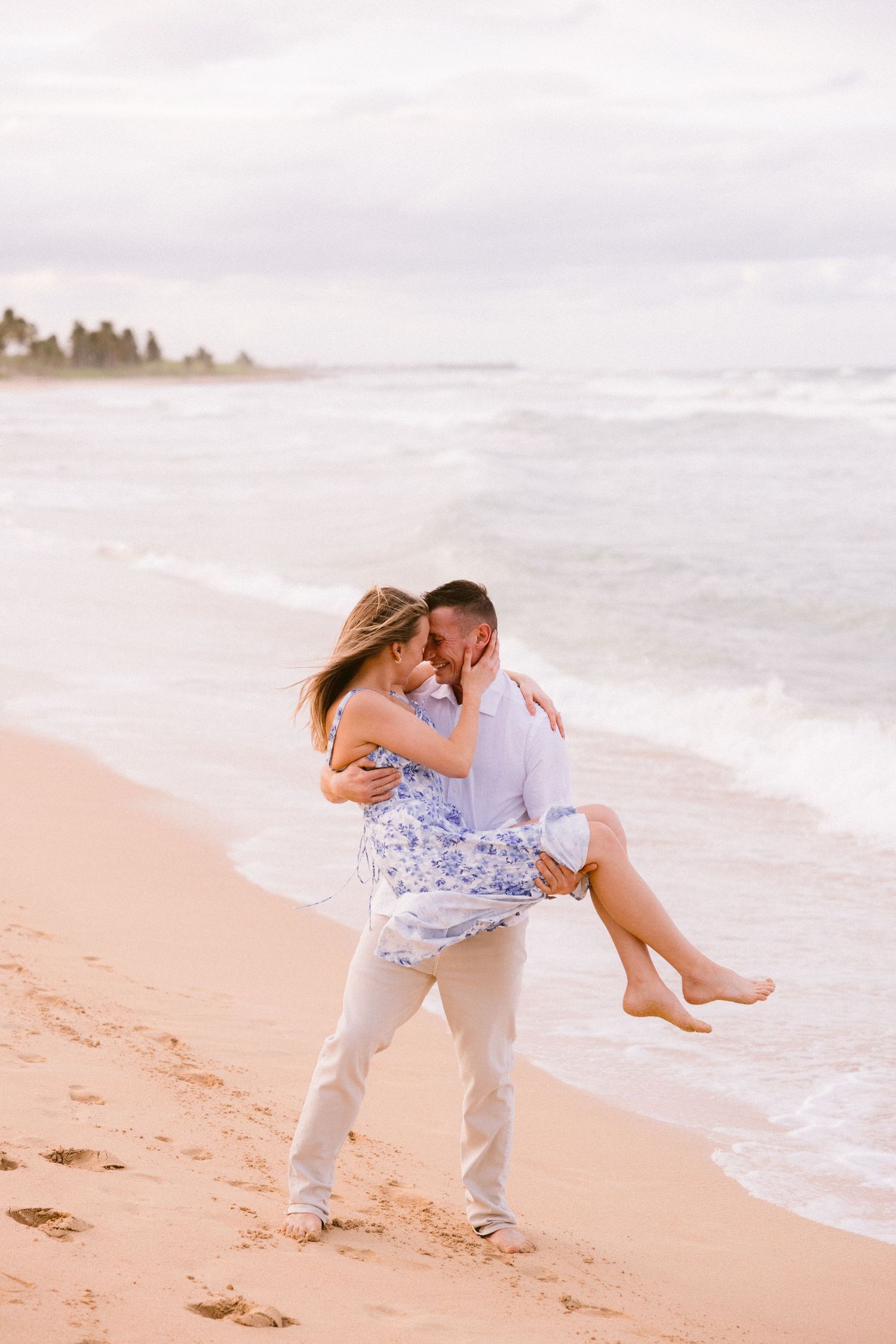 A romantic couple embraces on a sandy beach, man lifting woman in blue floral dress near ocean waves.