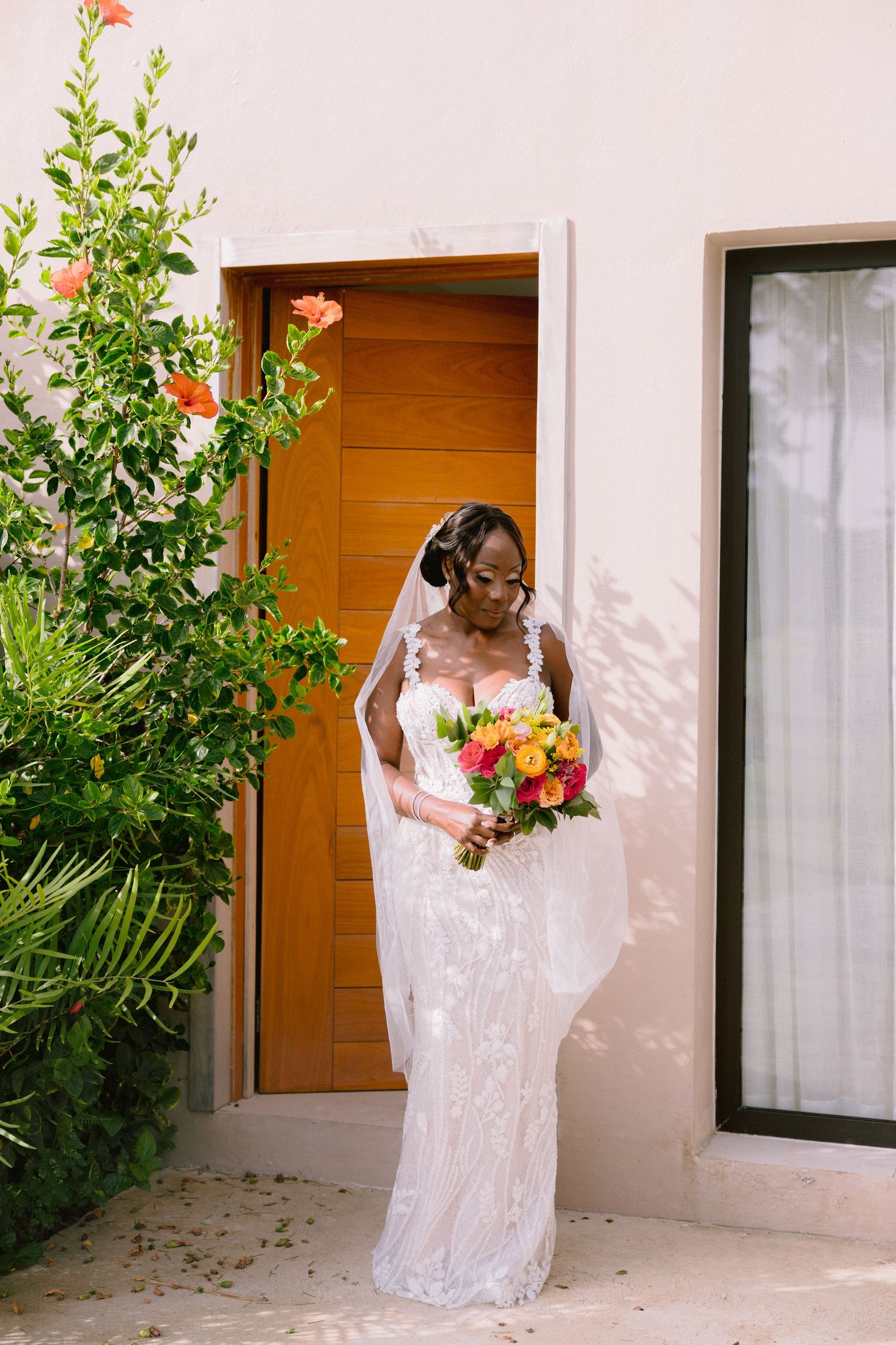A bride in a white lace wedding dress holds a vibrant bouquet of flowers against a white wall and wooden door.