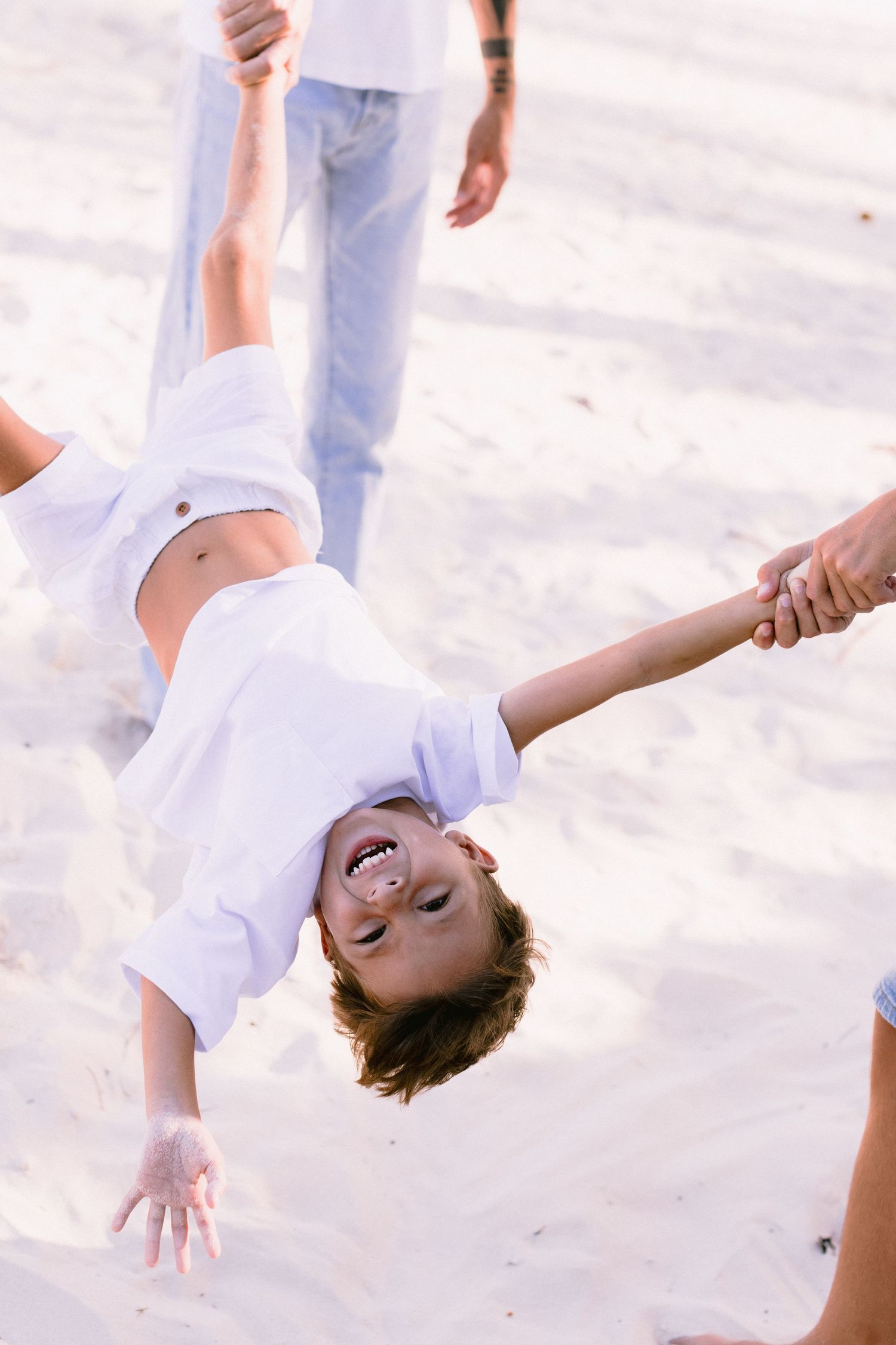 Joyful child being swung upside down by adults on a sandy beach, laughing with arms outstretched.