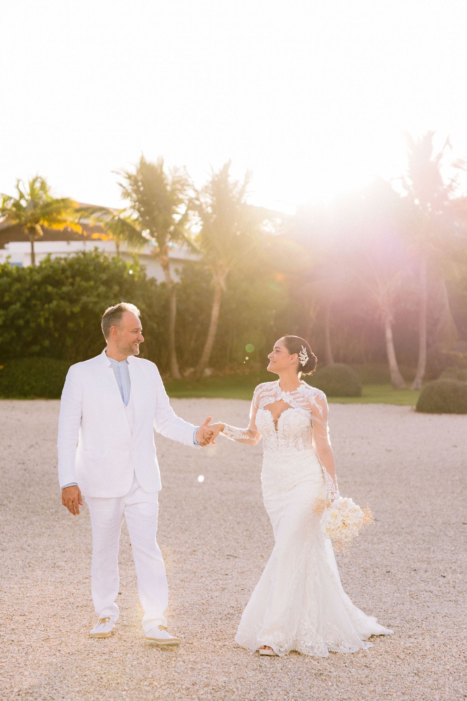 A couple in all white attire dances together on a sandy beach at sunset with palm trees in the background.