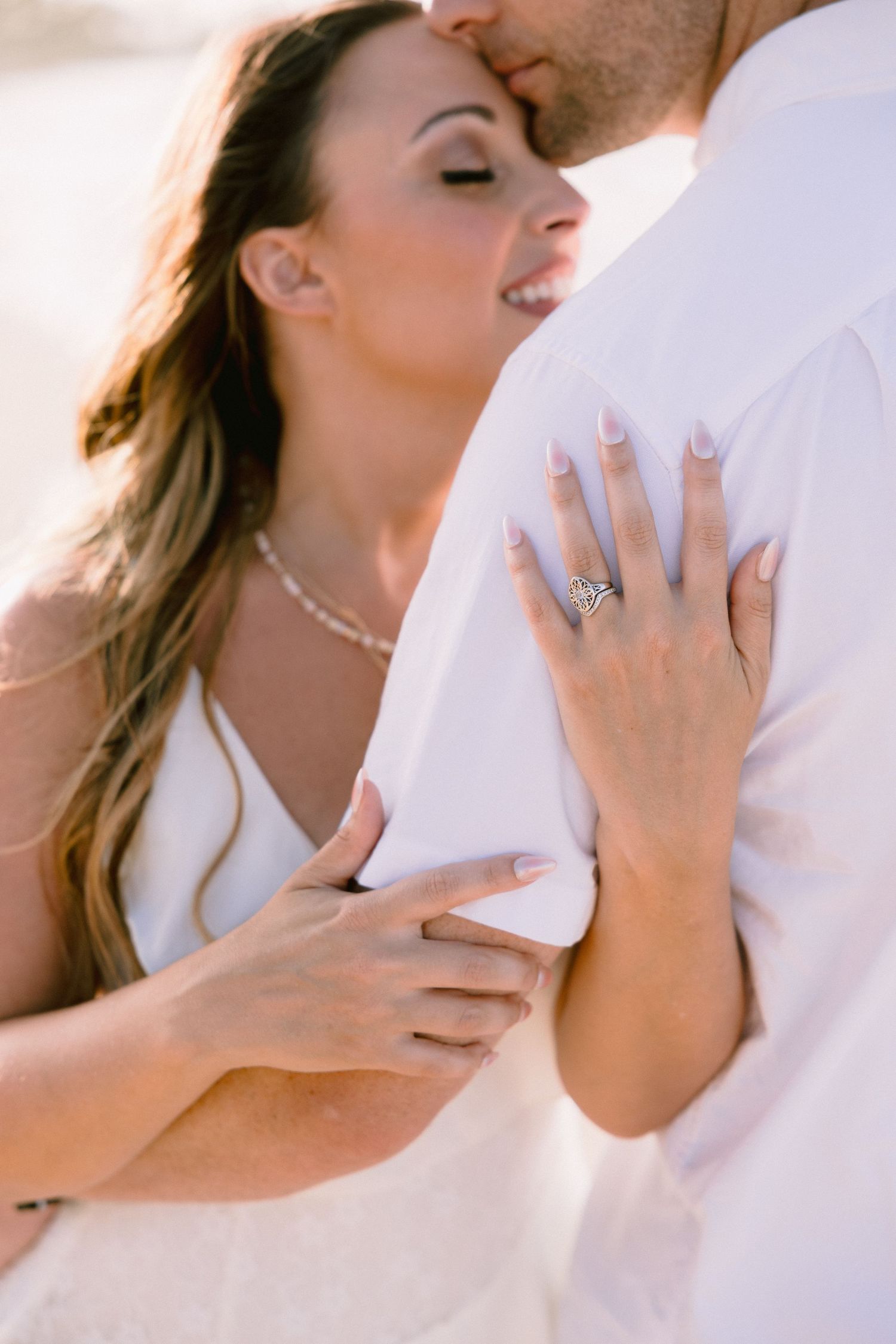 Couple in white embracing on beach, woman wearing diamond engagement ring and pearl necklace, romantic outdoor wedding photo.