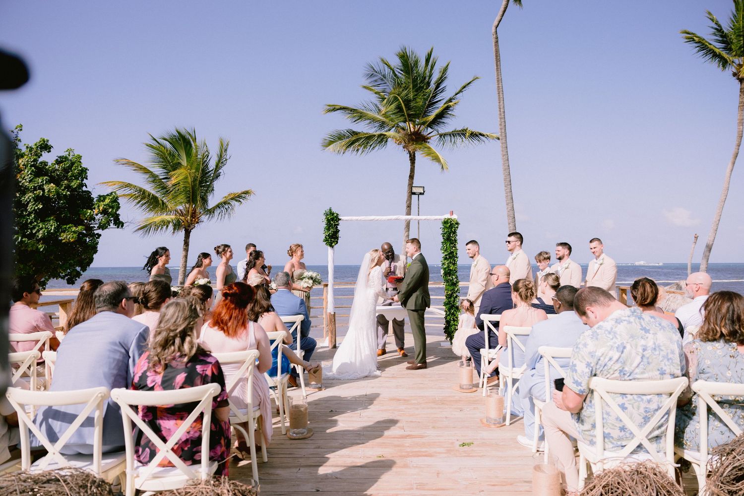 Outdoor beach wedding ceremony with guests seated in white chairs, palm trees, and ocean backdrop.