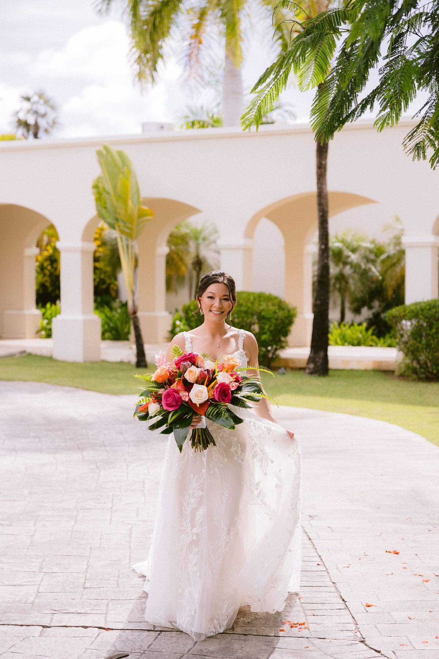 A bride in a white wedding dress sits elegantly on white chairs at an outdoor venue with arched columns and palm trees.