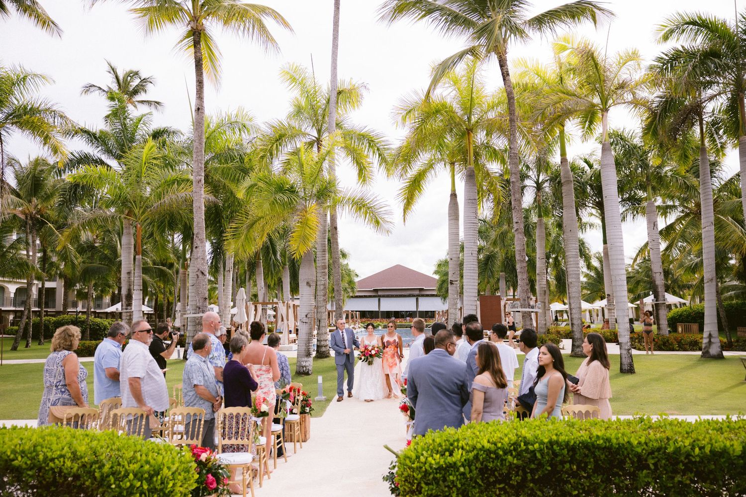 Wedding ceremony setup on a beach with white chairs and palm trees lining a sandy aisle in a tropical setting.