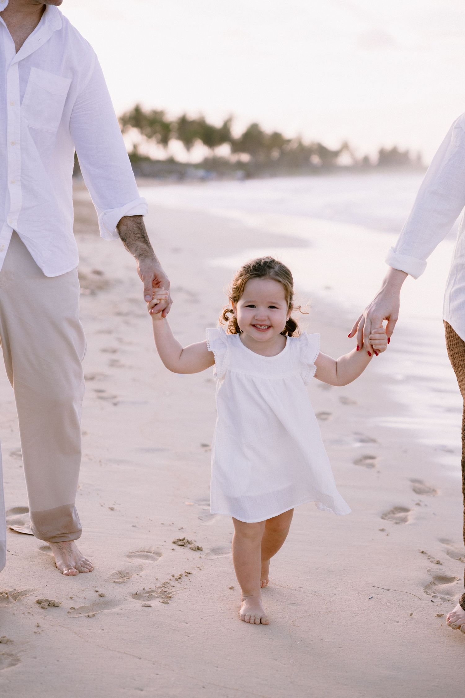 A small child in a white dress walks along a beach at sunset while holding hands with adults in white attire.