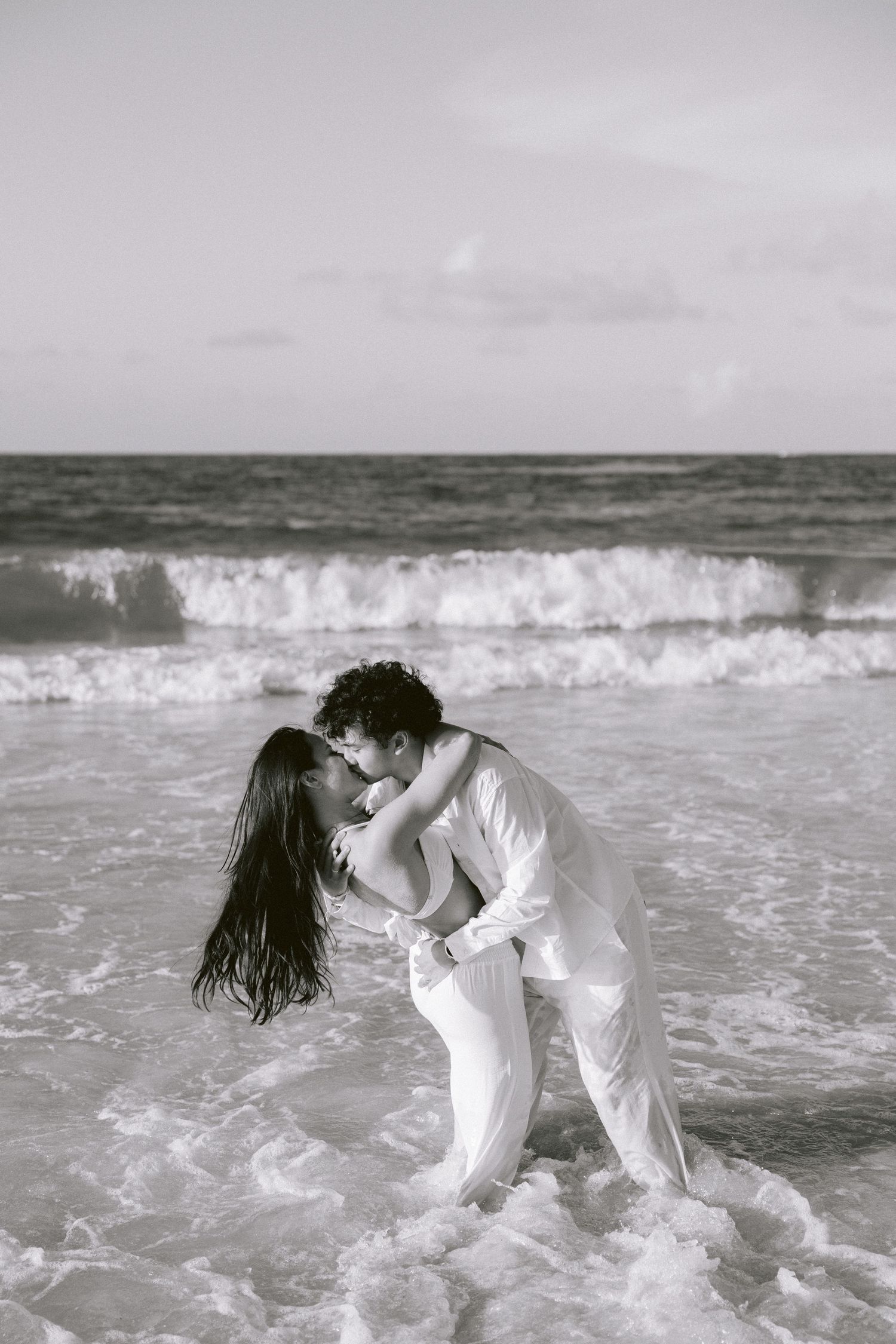 Couple sharing intimate moments in crashing ocean waves while wearing white clothing.