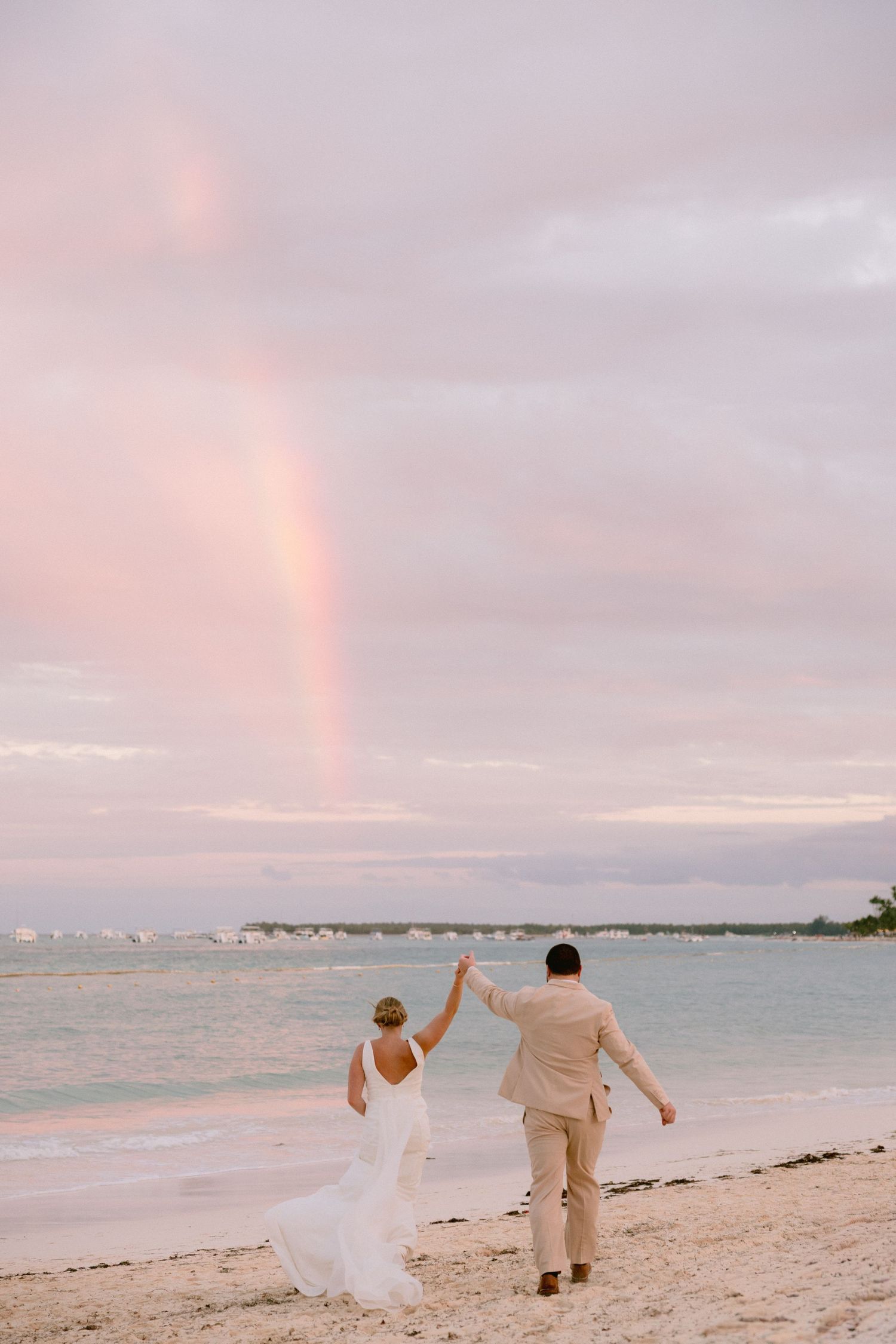 Bride and groom holding hands on a beach at sunset with a rainbow in the pink sky over the ocean.