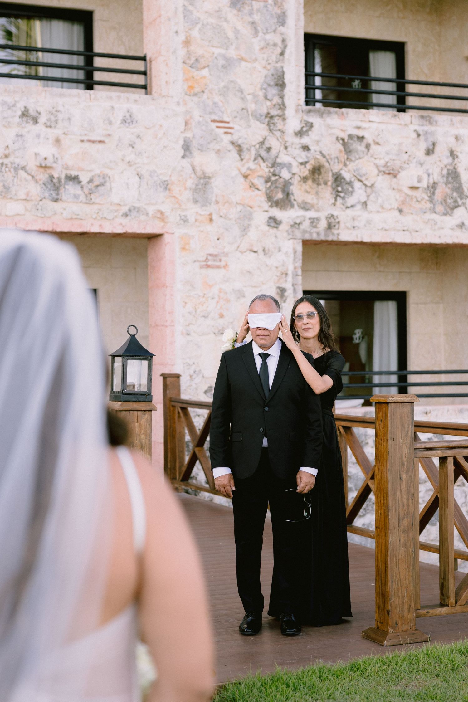 Blindfolded groom led by woman in black dress toward bride in white veil during wedding first look outdoors.