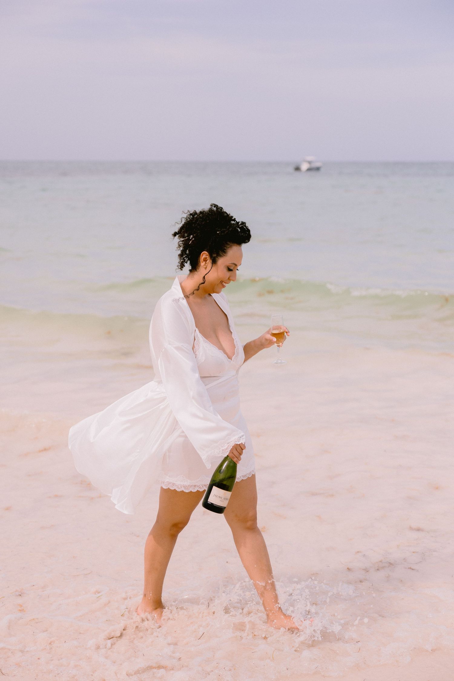 Woman in white dress walking on beach shore holding champagne bottle and glass with ocean in background.