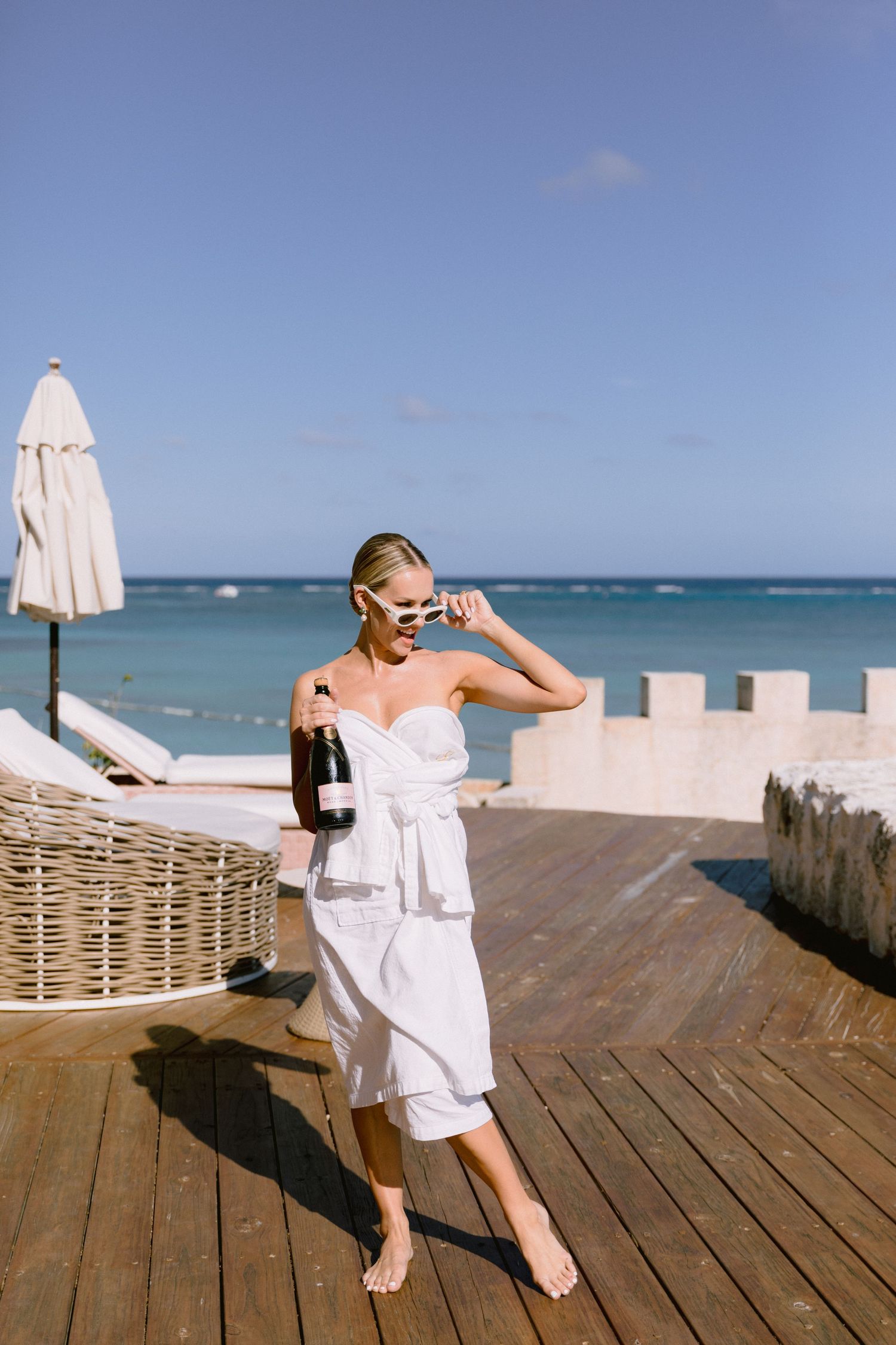 Woman in white wrap dress holding wine bottle on seaside deck with ocean view on sunny day.