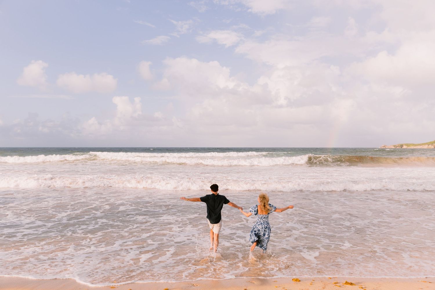 Two people running joyfully into the ocean waves on a sunny beach with white surf and blue sky.